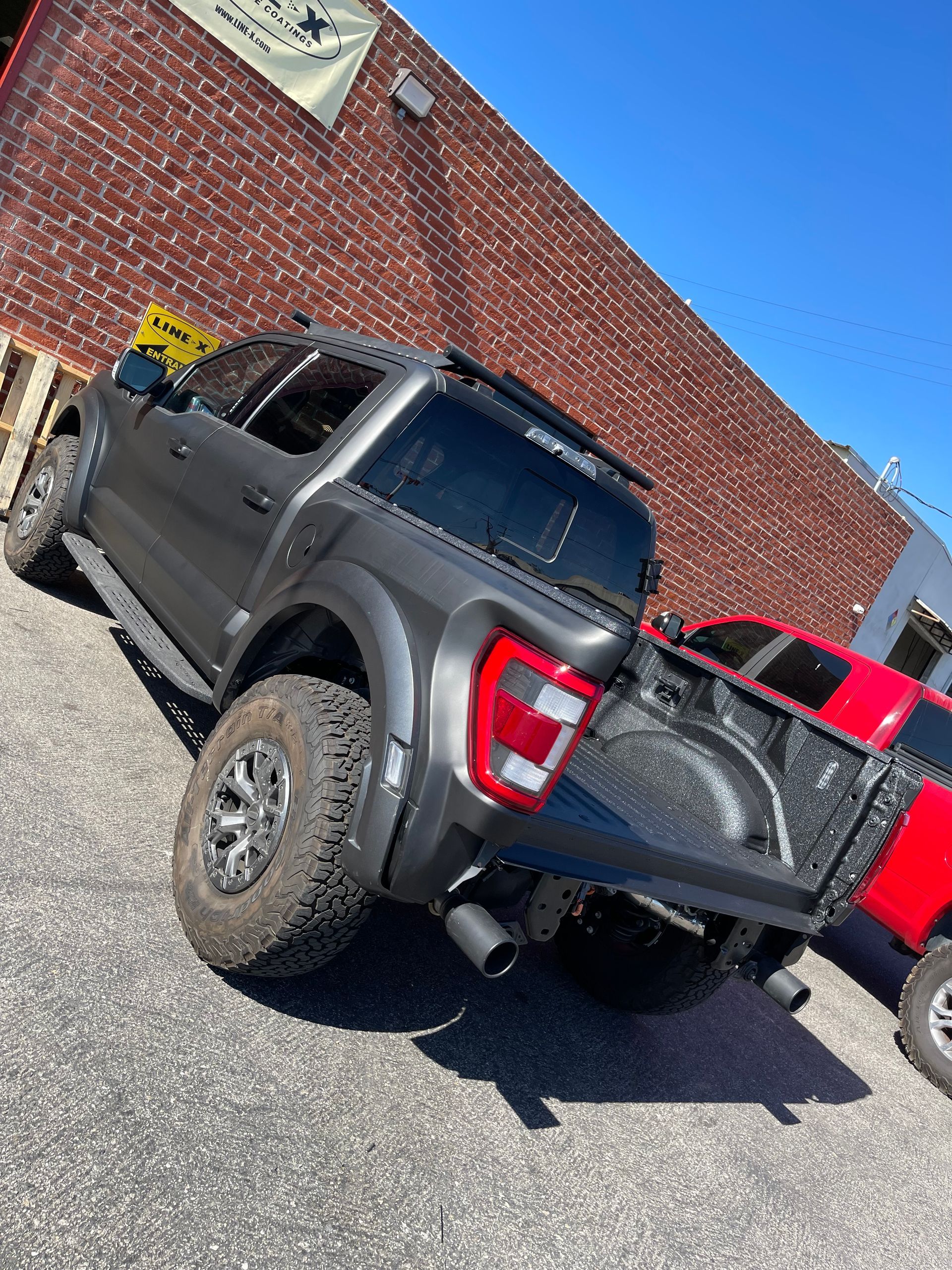 A matte dark grey pickup truck parked at an angle on an asphalt lot beside a red truck and a brick building.