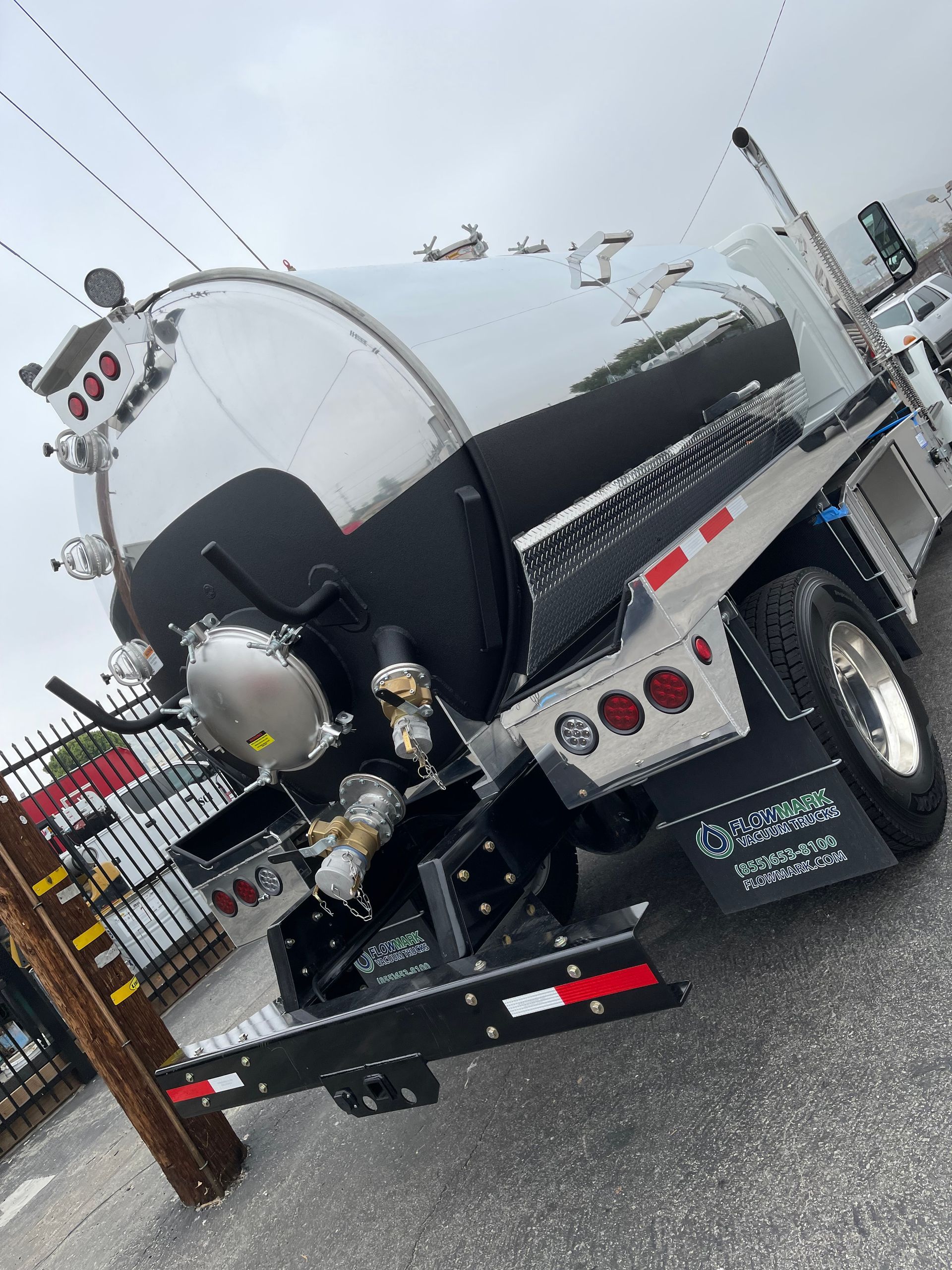 A chrome and black industrial vacuum tanker truck is parked on asphalt near a metal fence.