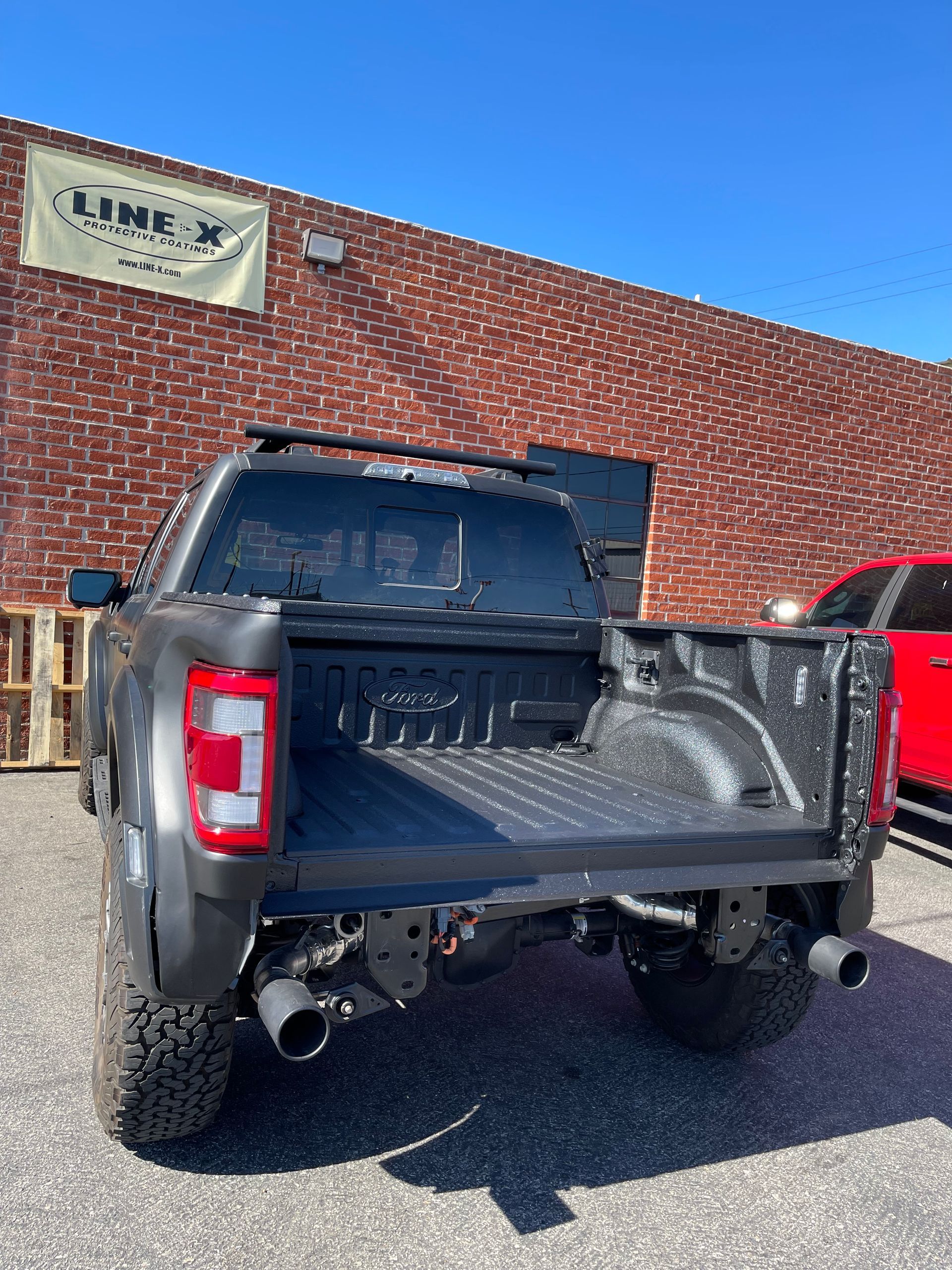 A dark-colored pickup truck with a spray-in bed liner parked in front of a brick building with a Line-X sign.