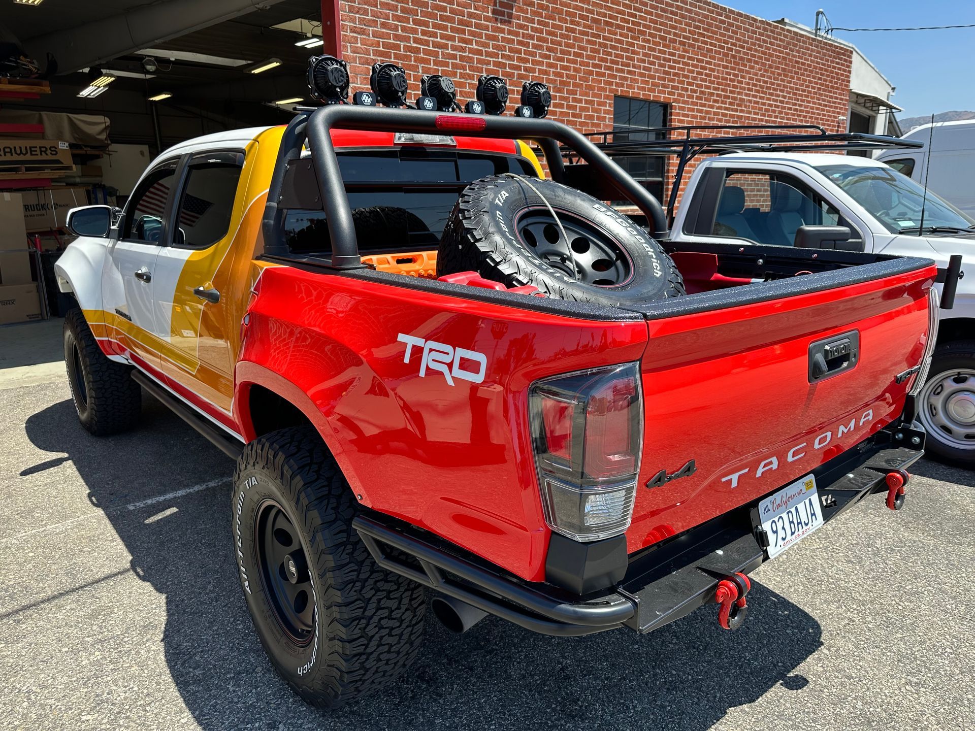 Red, white, and yellow Toyota Tacoma truck with a bed-mounted spare tire, light bar, and off-road bumper parked outside.