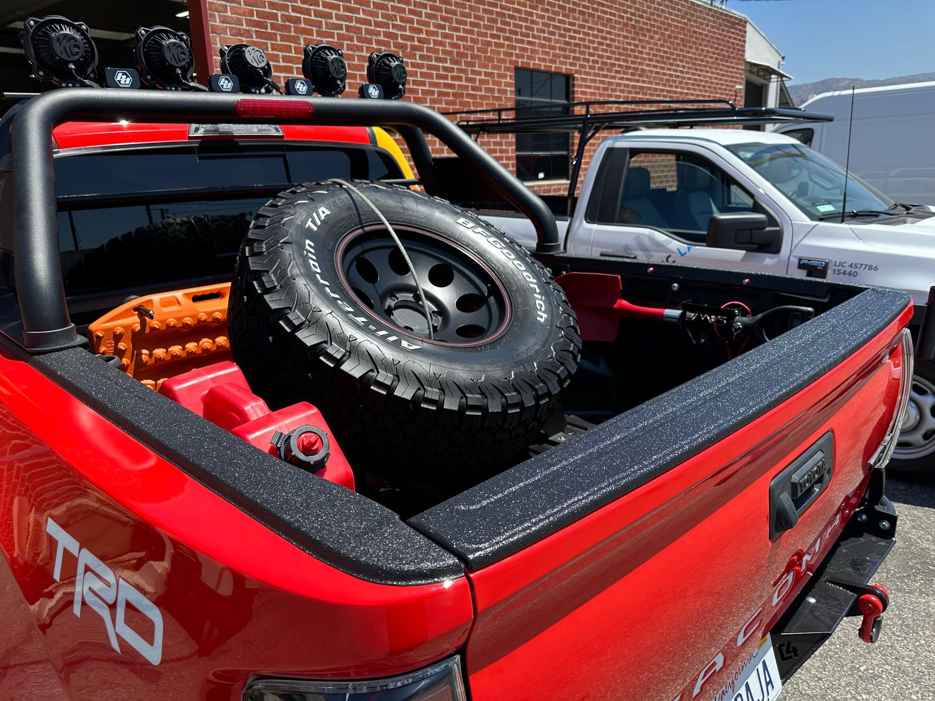 A red Toyota Tacoma truck bed with a spare tire, recovery boards, and a black roll bar with off-road lights.
