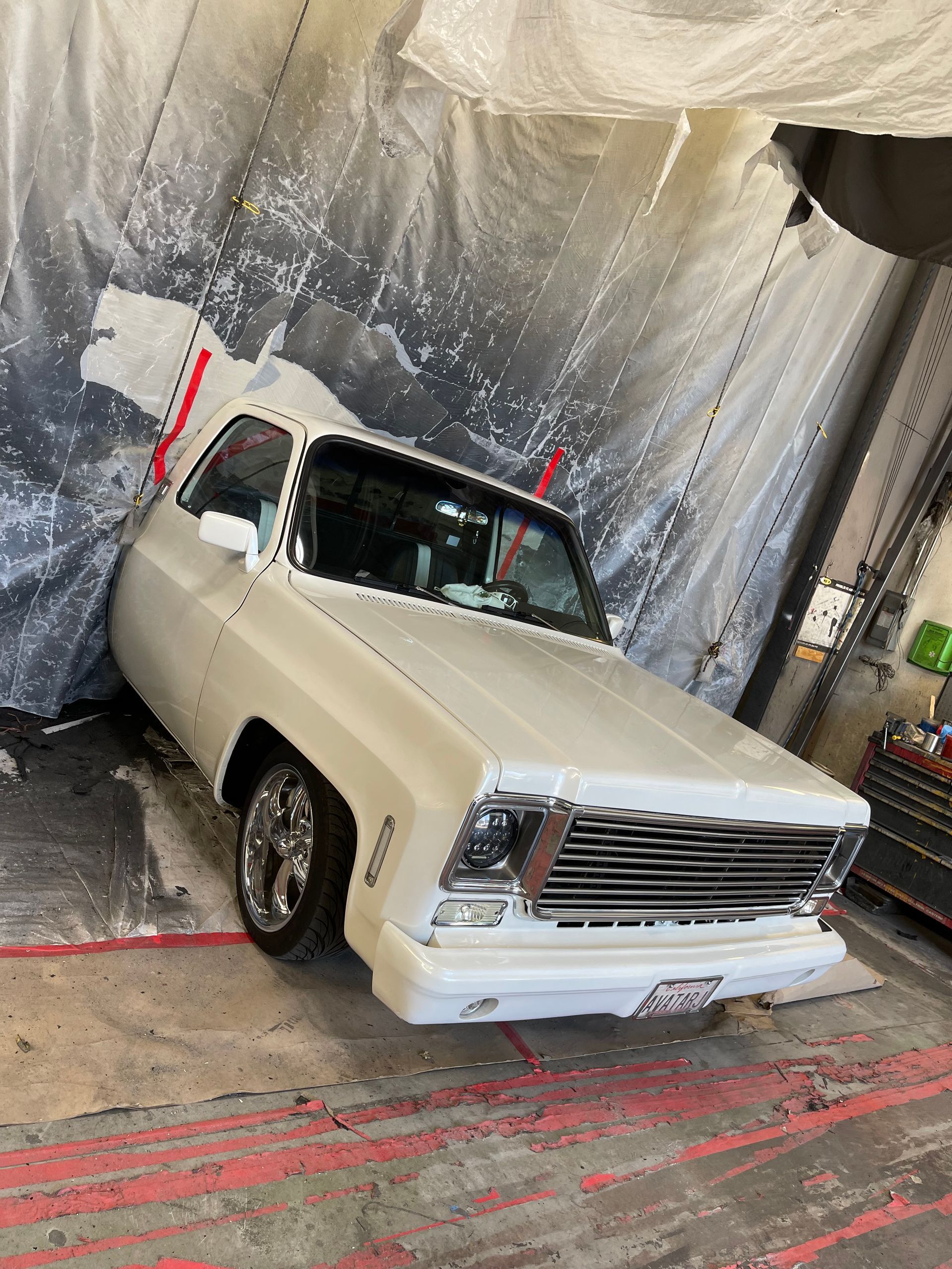A white classic pickup truck with chrome wheels parked inside a paint booth enclosed by plastic sheeting.