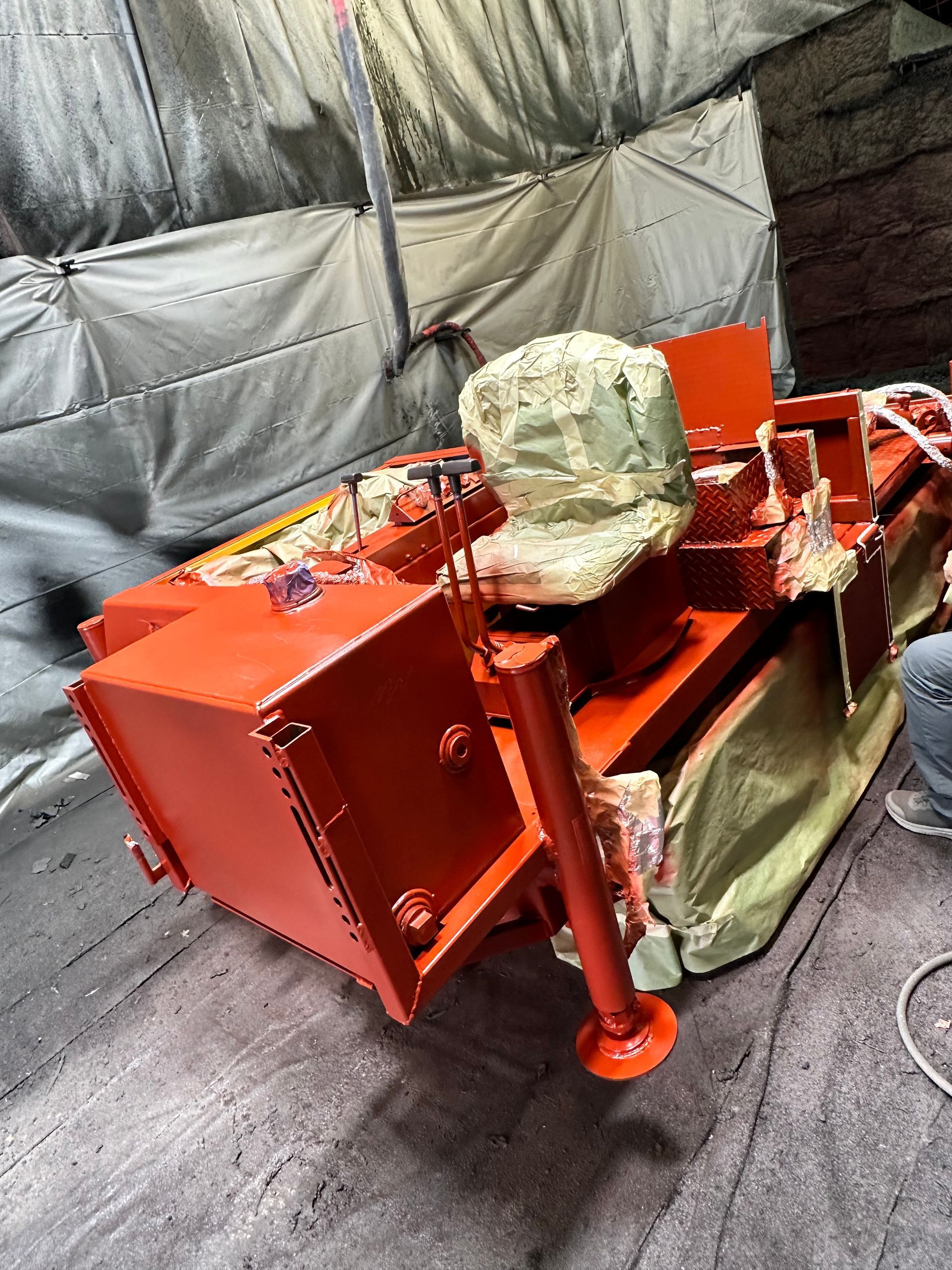 A bright orange industrial vehicle, partially covered by protective plastic sheets, sits in an indoor workspace.