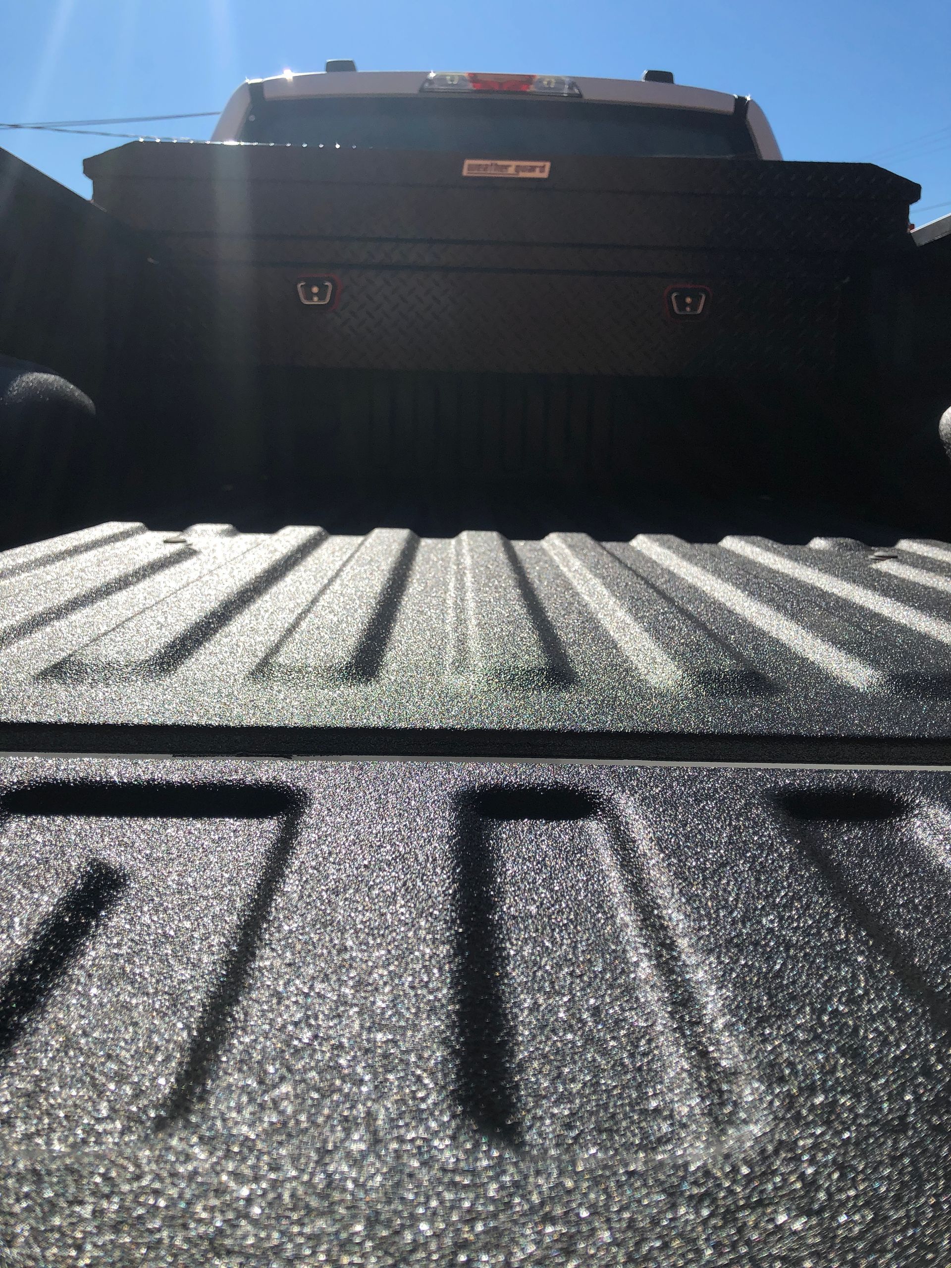 A black pickup truck bed with a textured spray-in liner and a dark metal toolbox against the back window.
