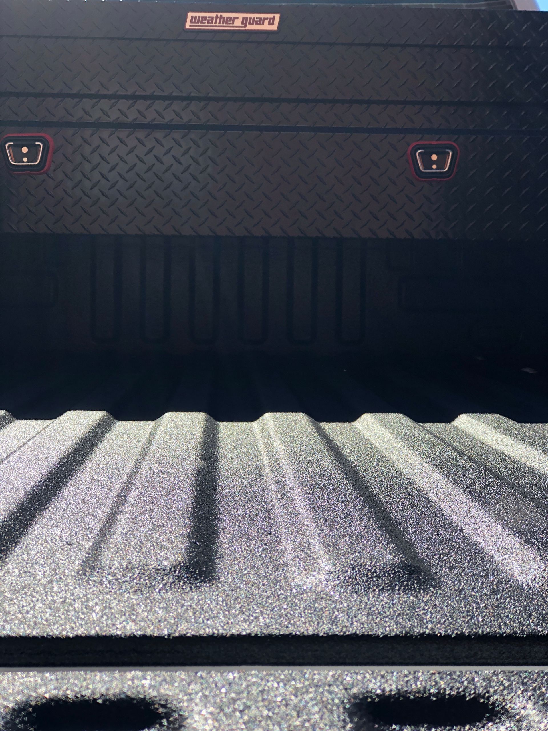 A black diamond-plate truck tool box is mounted at the front of a truck bed with a textured black spray-in bed liner.