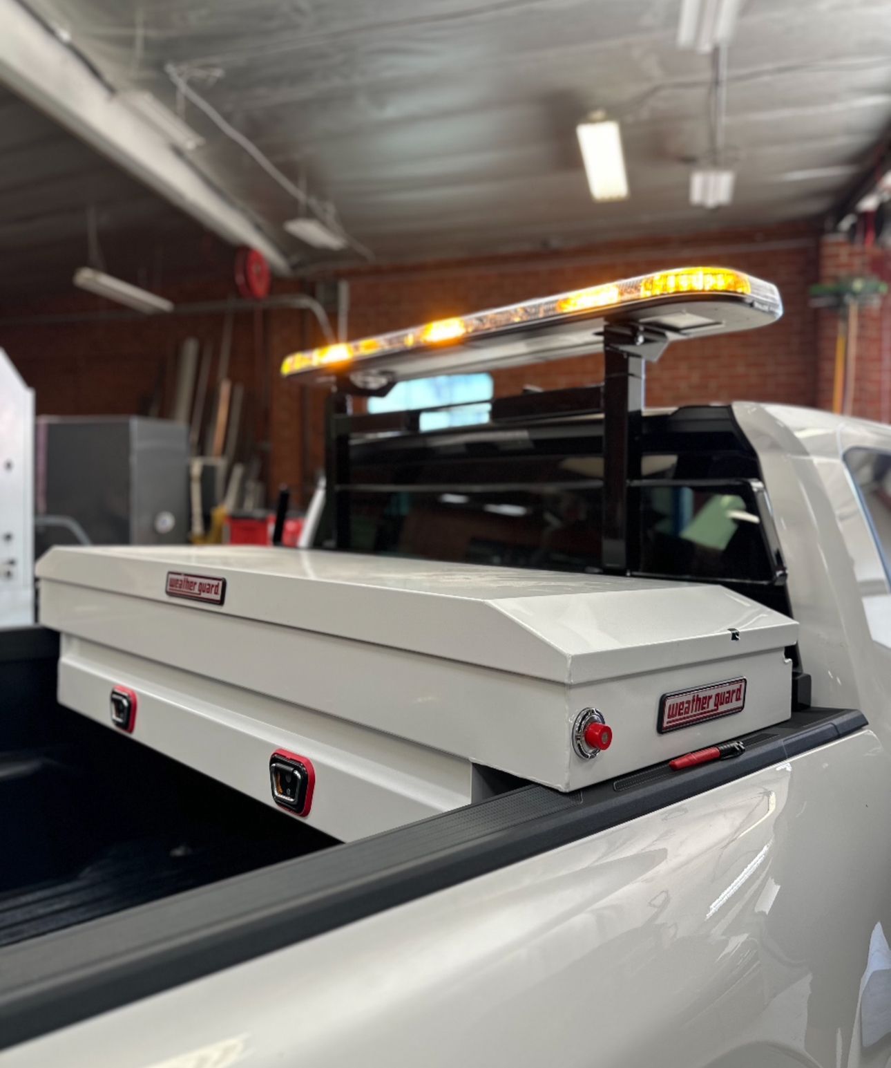 A white pickup truck bed featuring a matching white storage toolbox and a mounted amber emergency light bar.