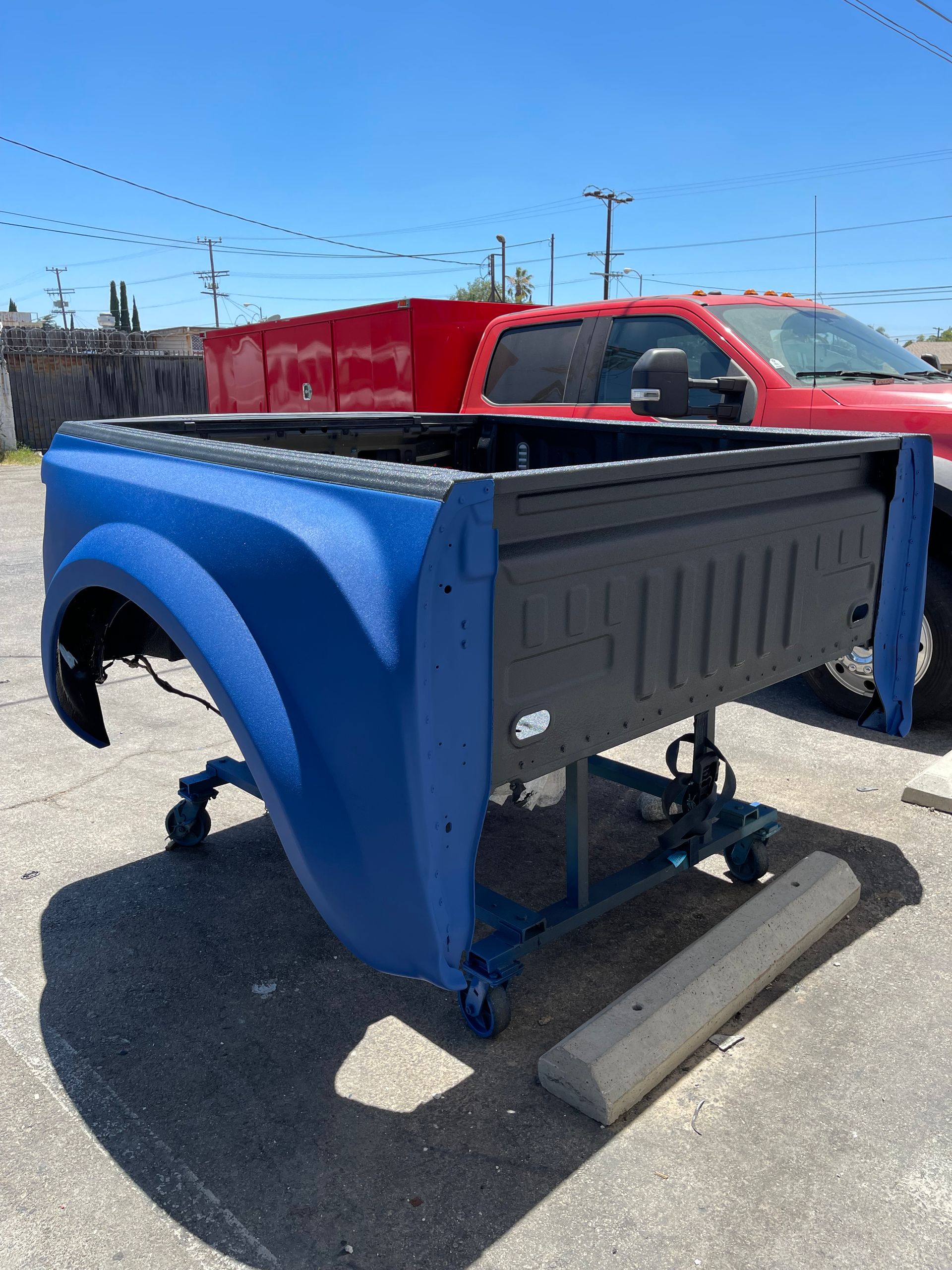 A blue pickup truck bed on a metal rolling stand, situated in a paved outdoor lot with a red truck in the background.