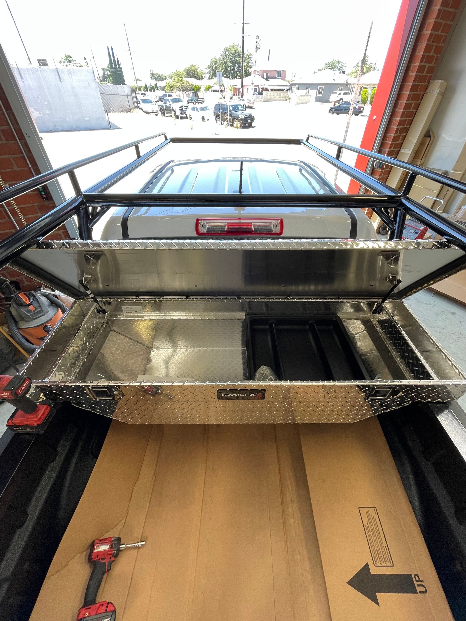 An open silver aluminum tool box sits in the bed of a white pickup truck, seen from above with a drill on the bed floor.