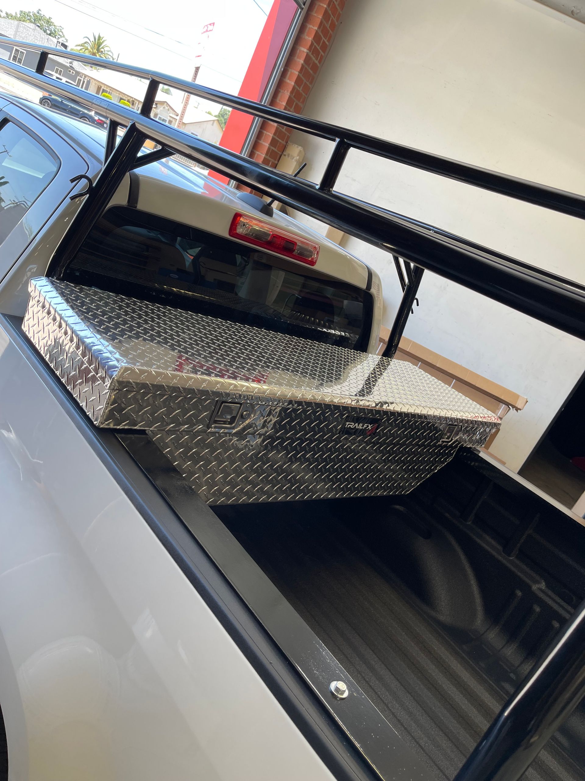 A metal diamond-plate tool box sits mounted in the bed of a white pickup truck, beneath a black metal ladder rack.