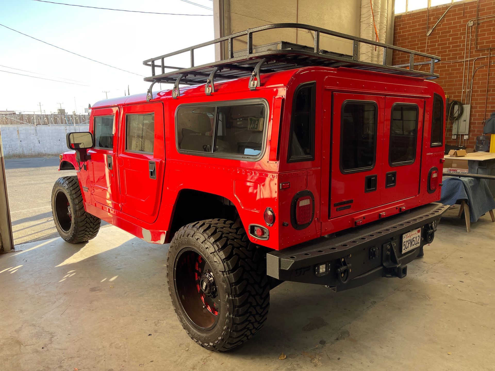A bright red, modified Hummer H1 parked in a garage, featuring large off-road tires, a rear roof rack, and tinted windows.