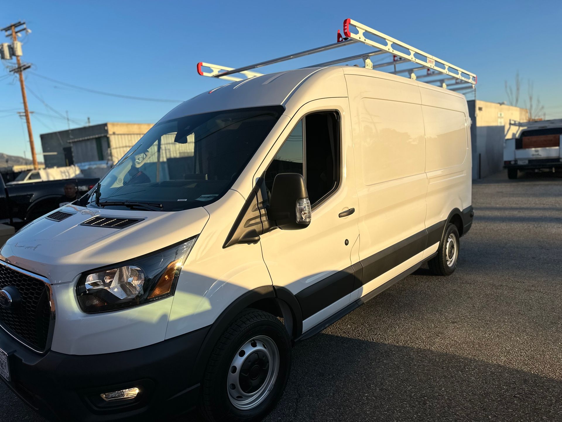 A white Ford Transit cargo van parked on a gravel lot with a utility roof rack installed on top.