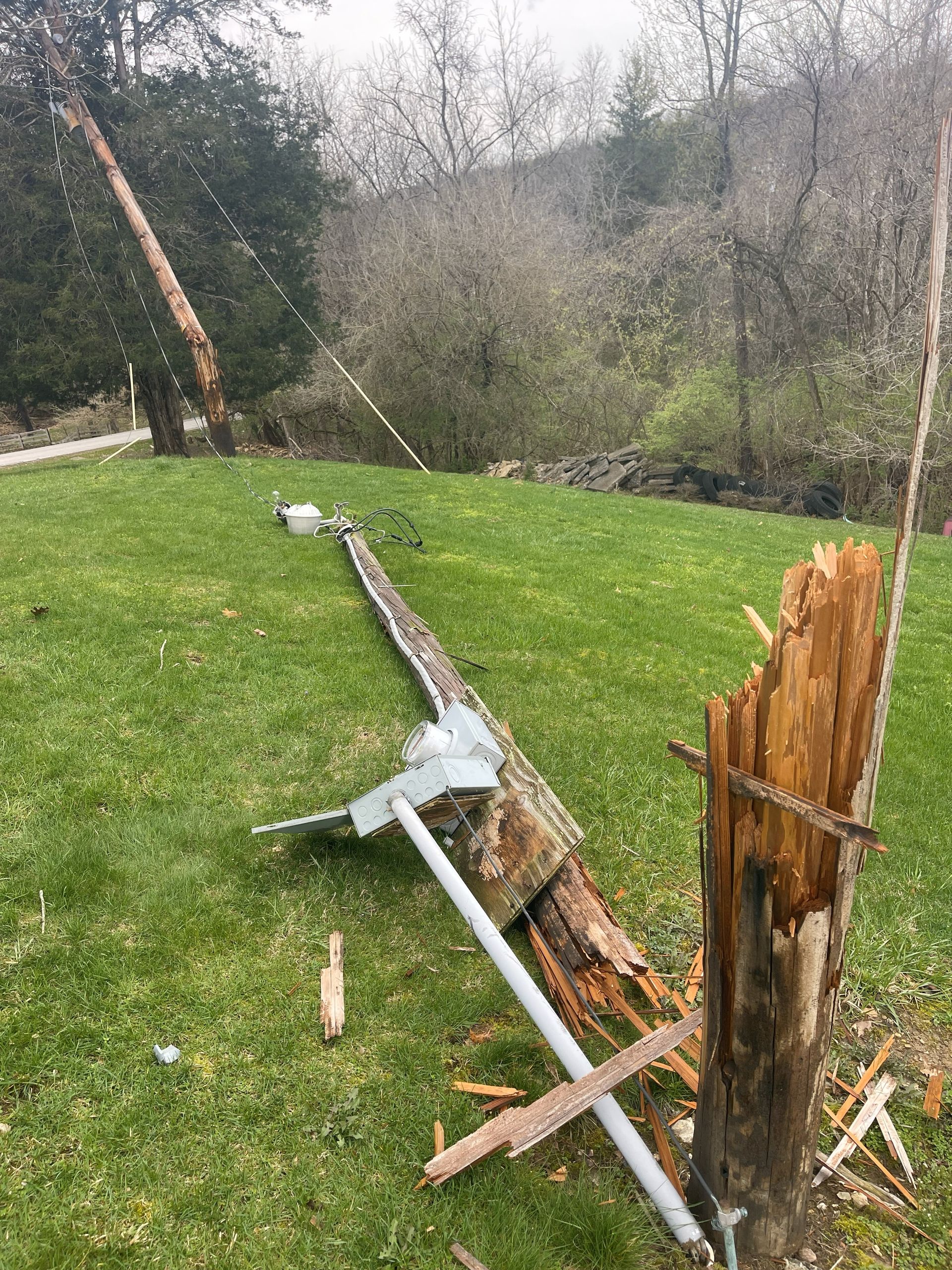 A broken utility pole lies on a grassy lawn; wires are still attached, and wood is splintered.