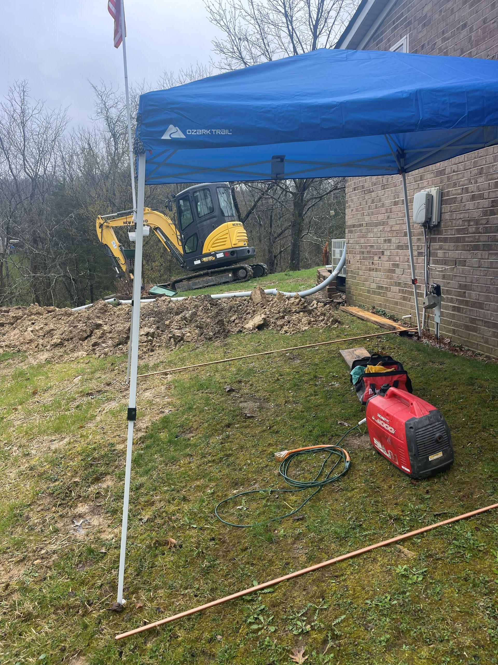 Construction site: Excavator digging. Blue canopy, generator, tools on grass near brick building.