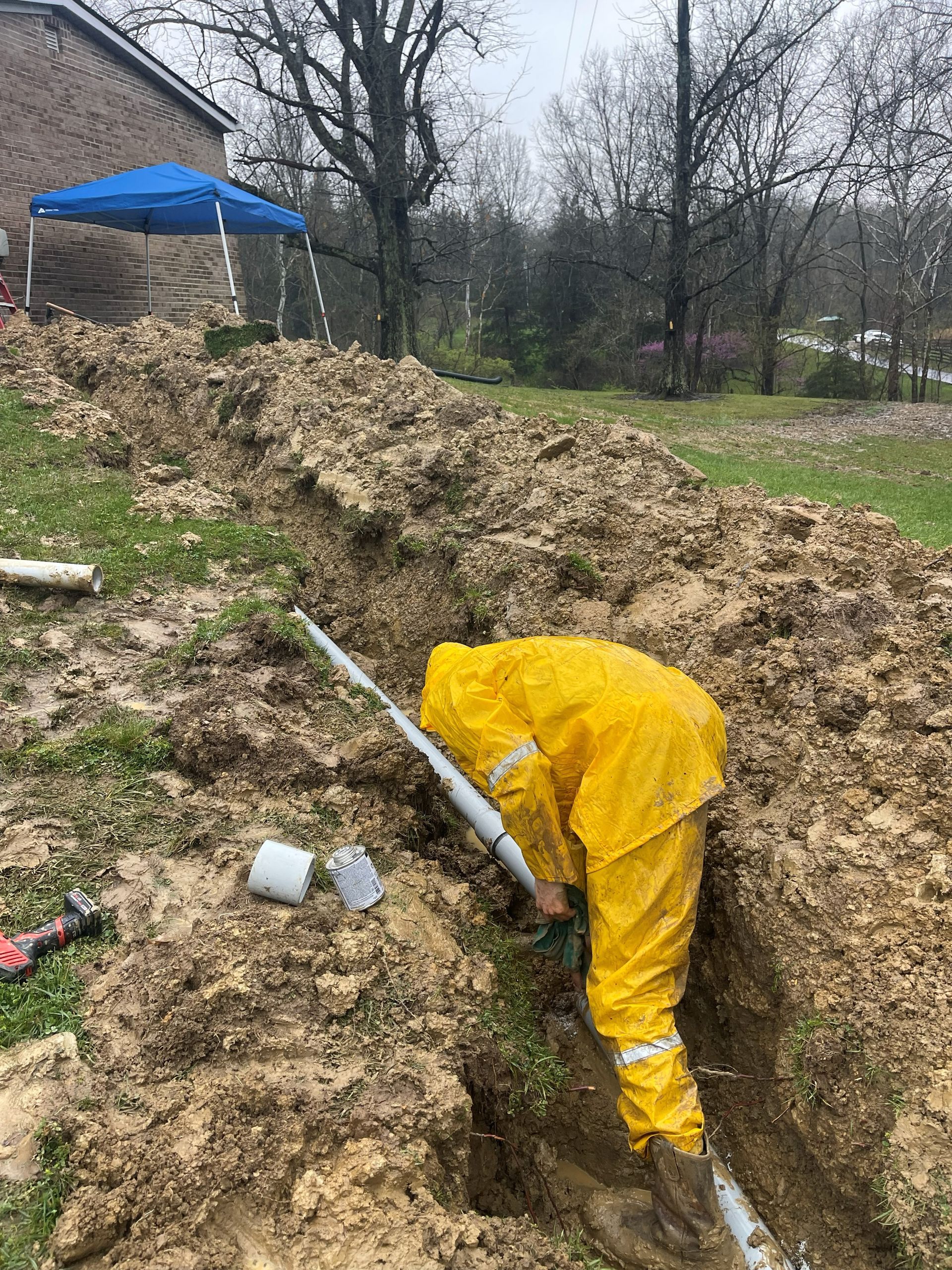 Person in yellow protective suit working on pipes in a trench outdoors near a house.