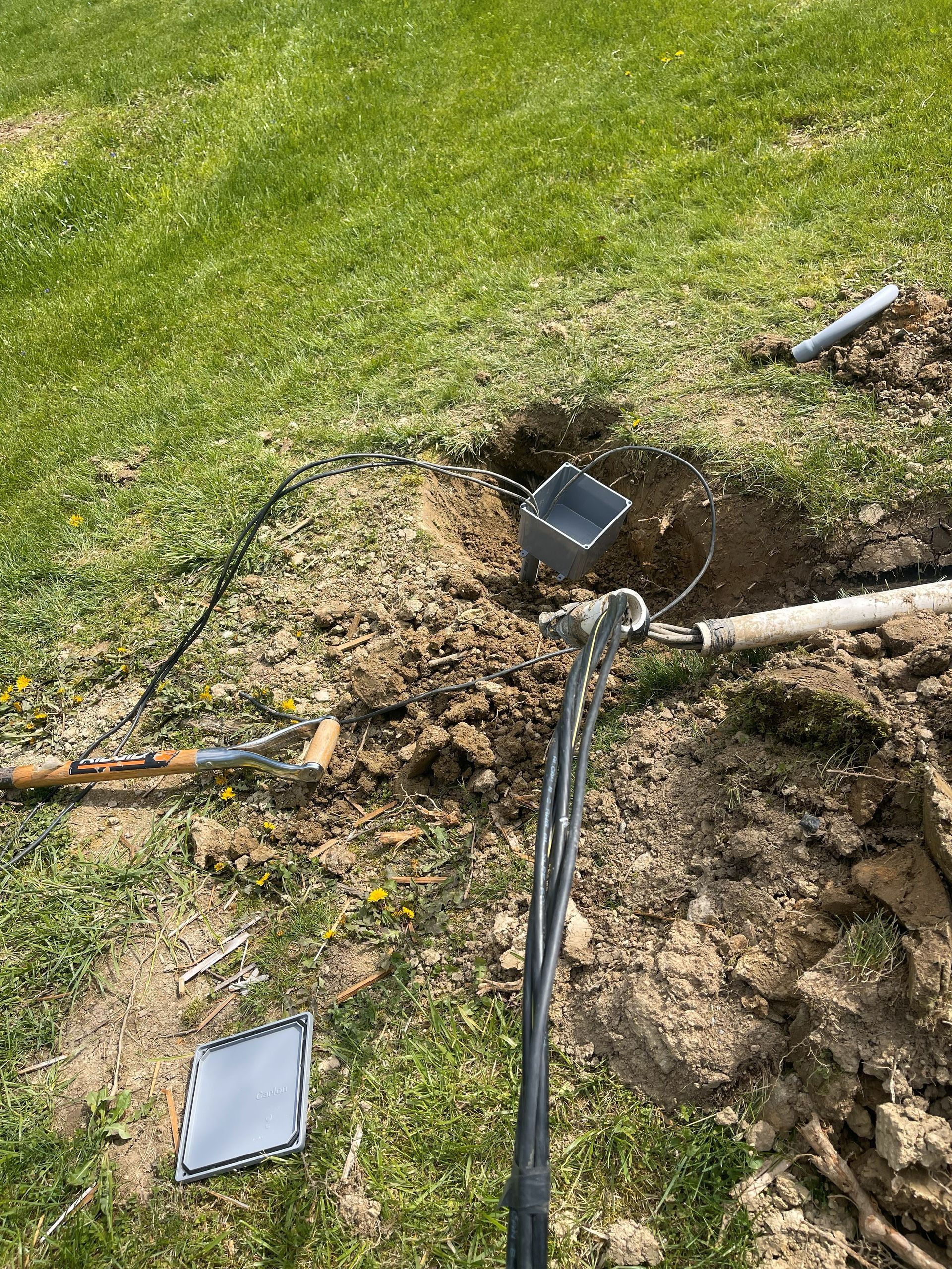 Hole in grass with exposed electrical wires, metal box, and tools. Dirt and pipes visible.