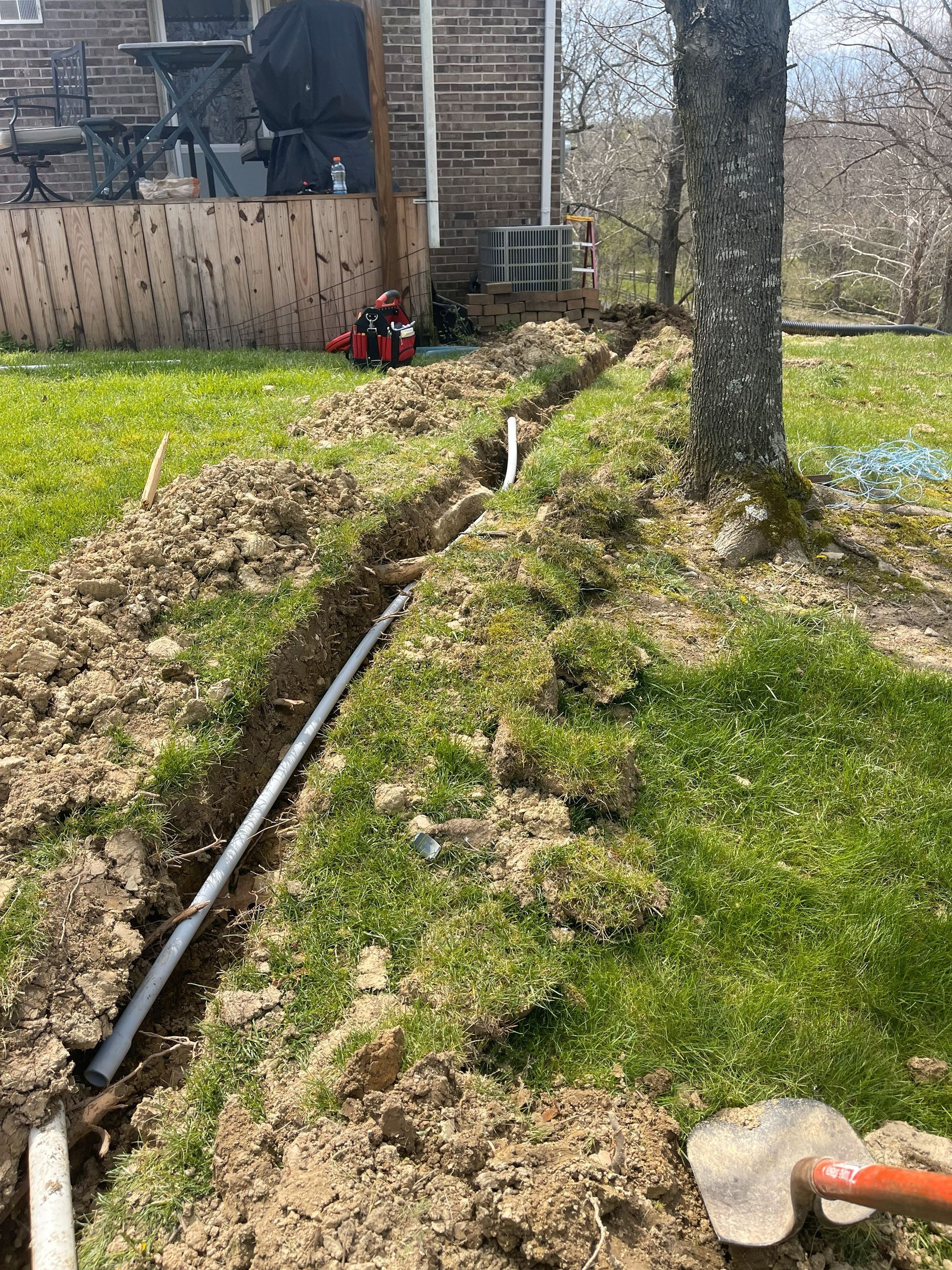 Trench dug in a grassy yard, containing a gray pipe. A tree and house are in the background. A shovel rests nearby.