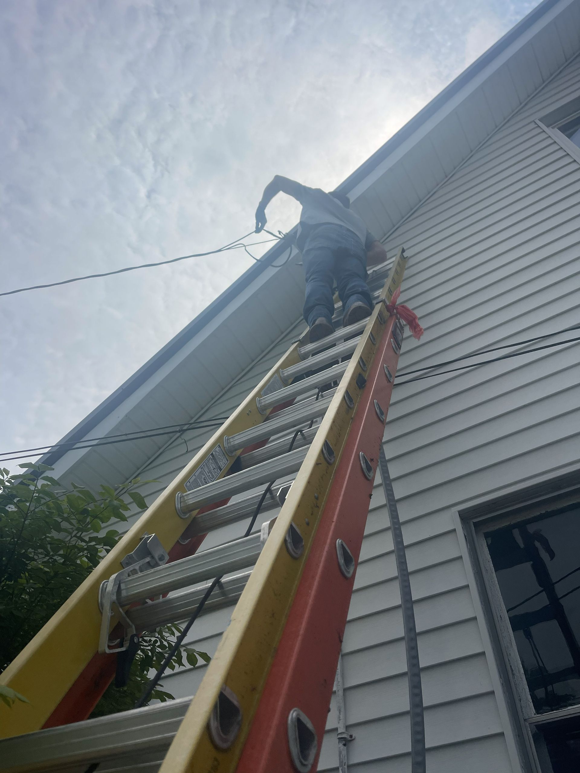 Person on ladder working on wires attached to a white house with a blue trim. Overcast sky in the background.