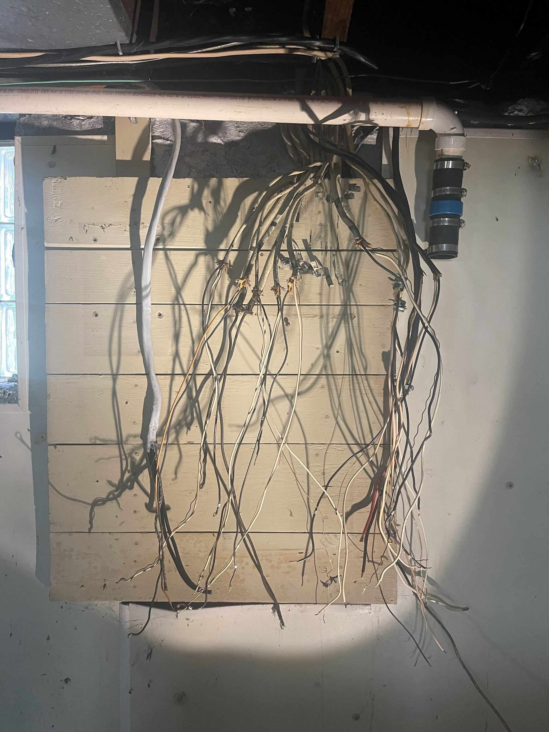 Exposed electrical wires hanging from a wooden panel in a basement.