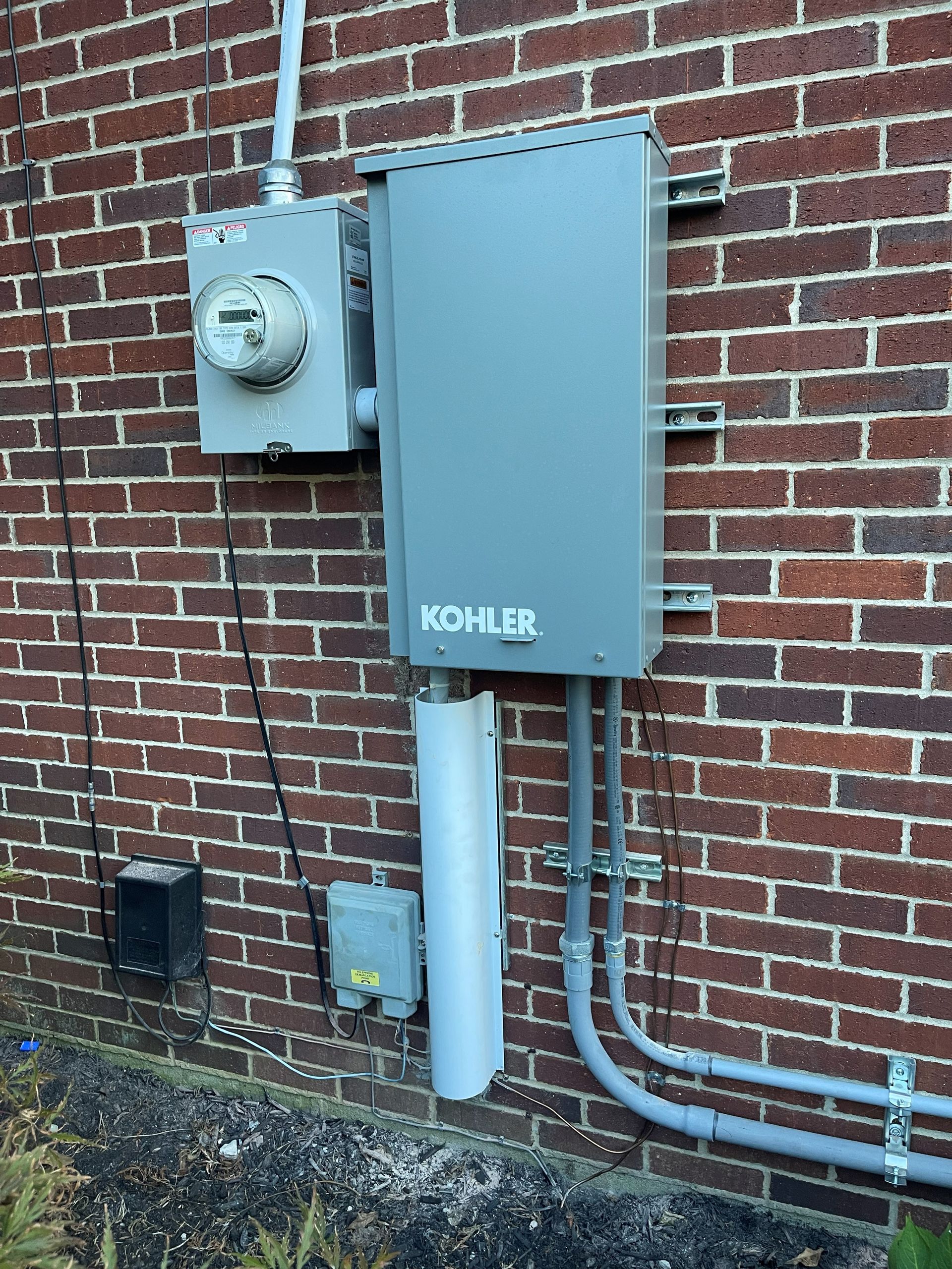 Exterior electrical equipment on a brick wall: meter, power transfer switch, conduits, and a Kohler brand generator enclosure.