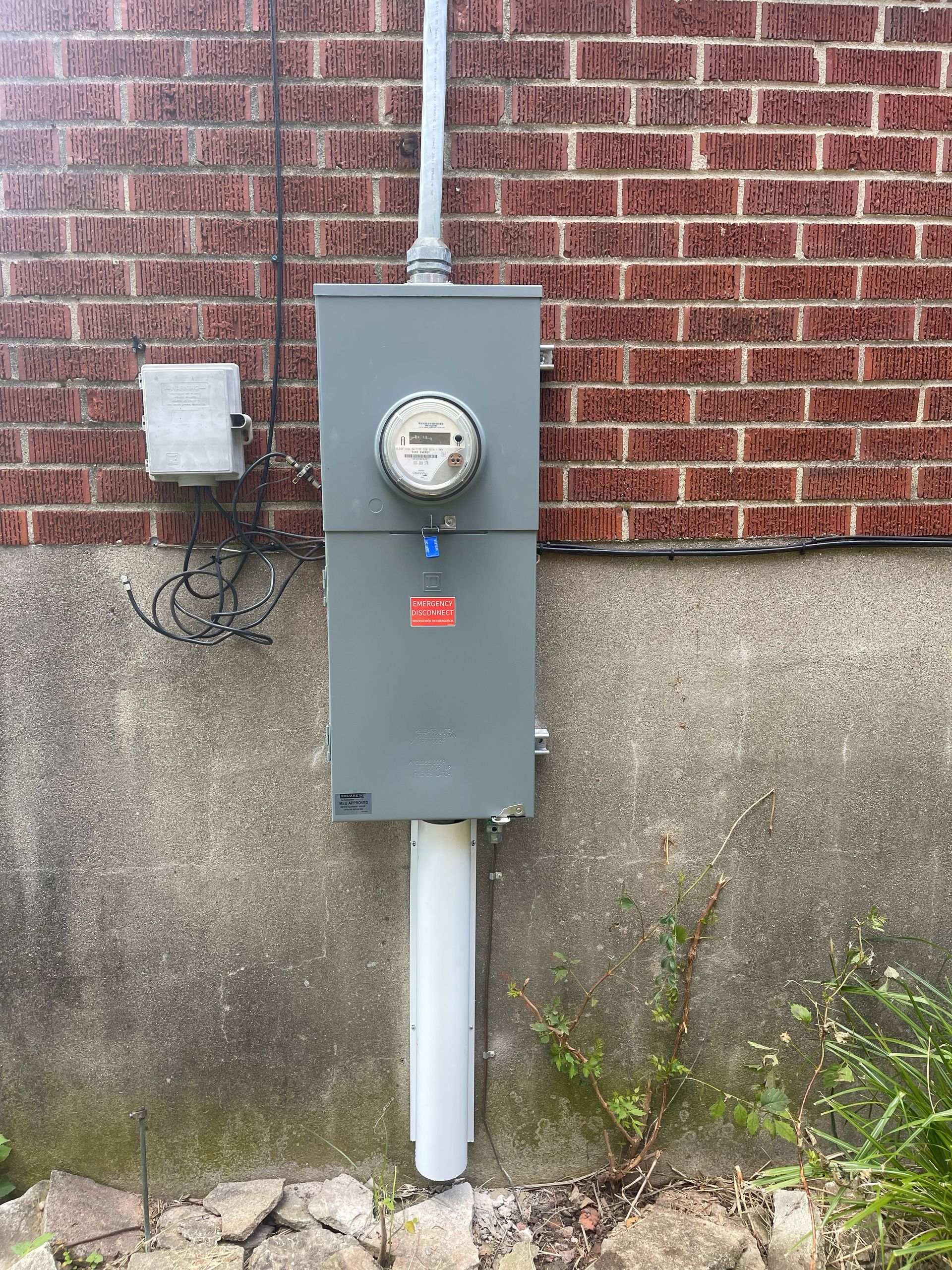 Electric meter box mounted on a brick and concrete wall. Gray metal box, white conduit, digital meter.