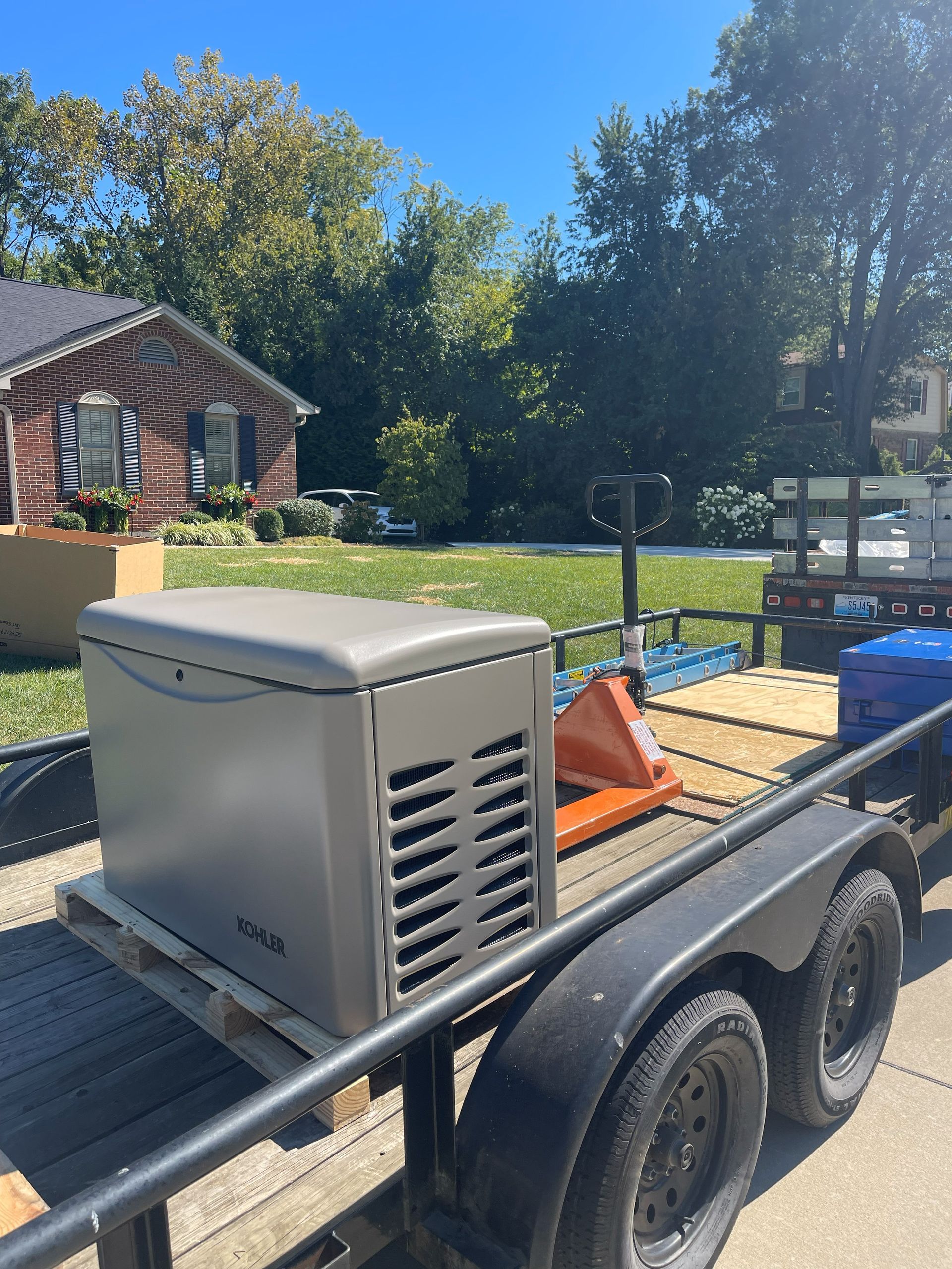 Generator on a trailer in front of a house.  The generator is gray.  Blue skies and trees are in the background.