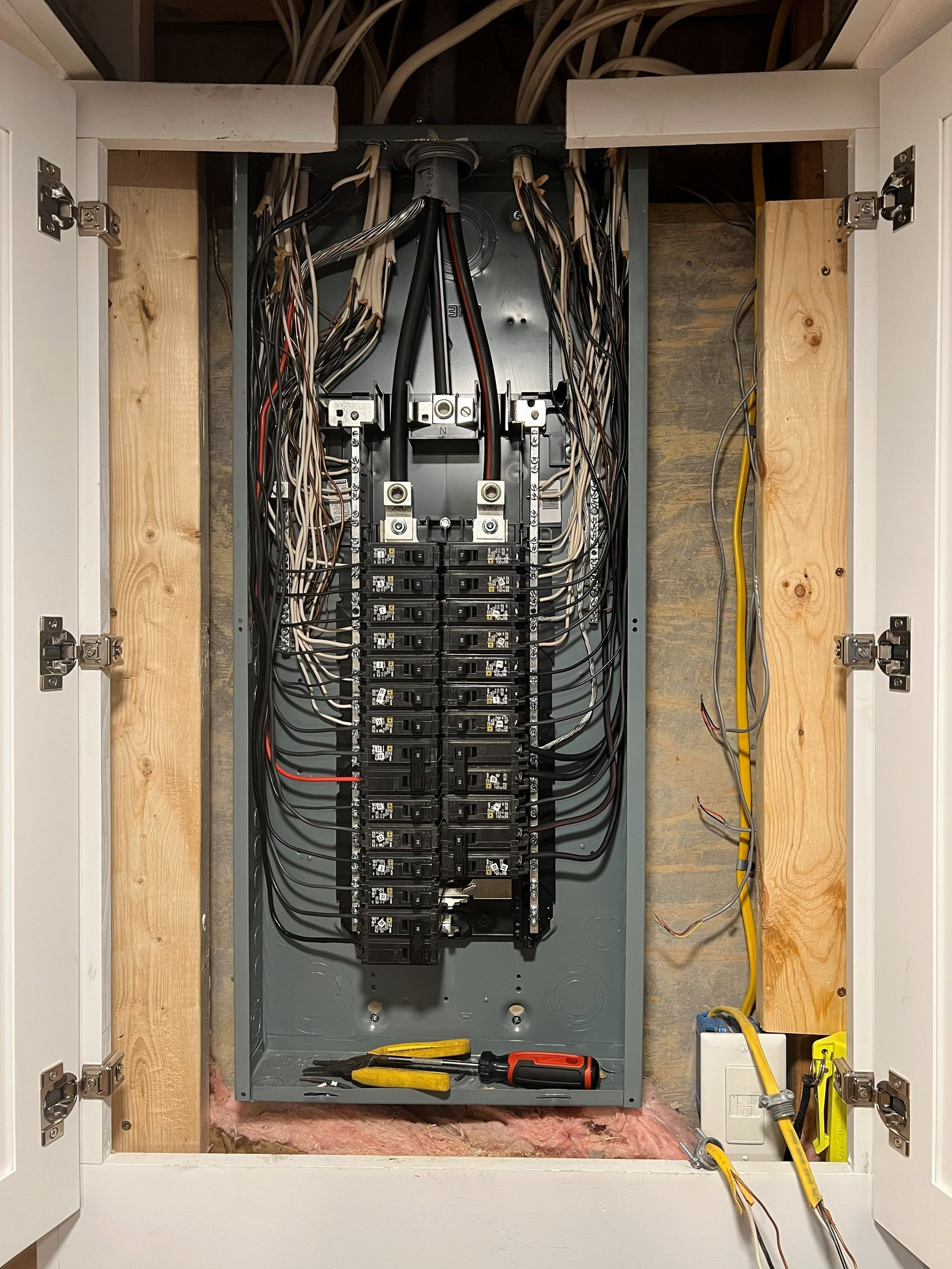 Electrical panel with exposed wiring inside a cabinet with open white doors, tools visible.