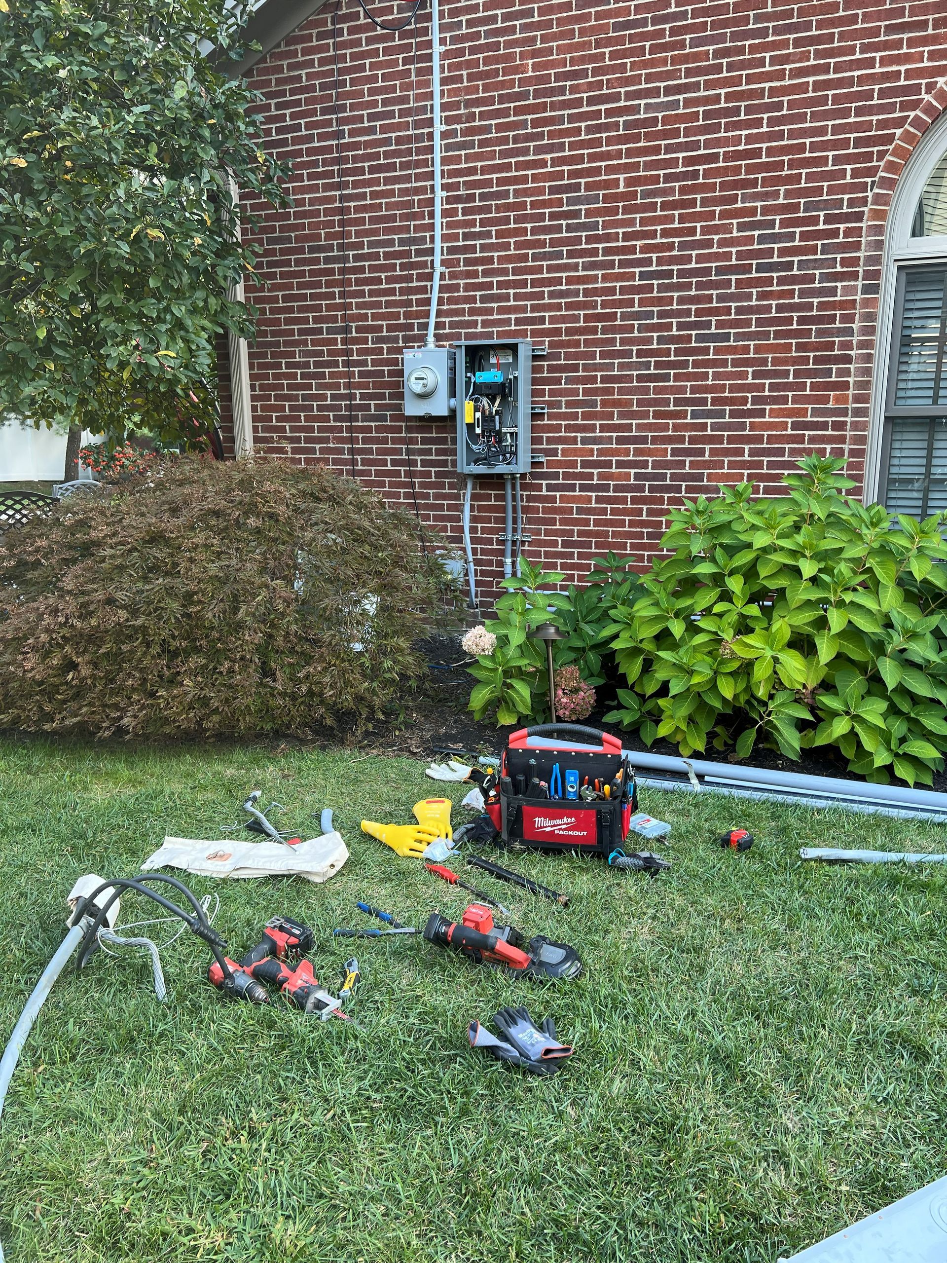 Electrical tools on grass in front of a brick building with an electrical panel.