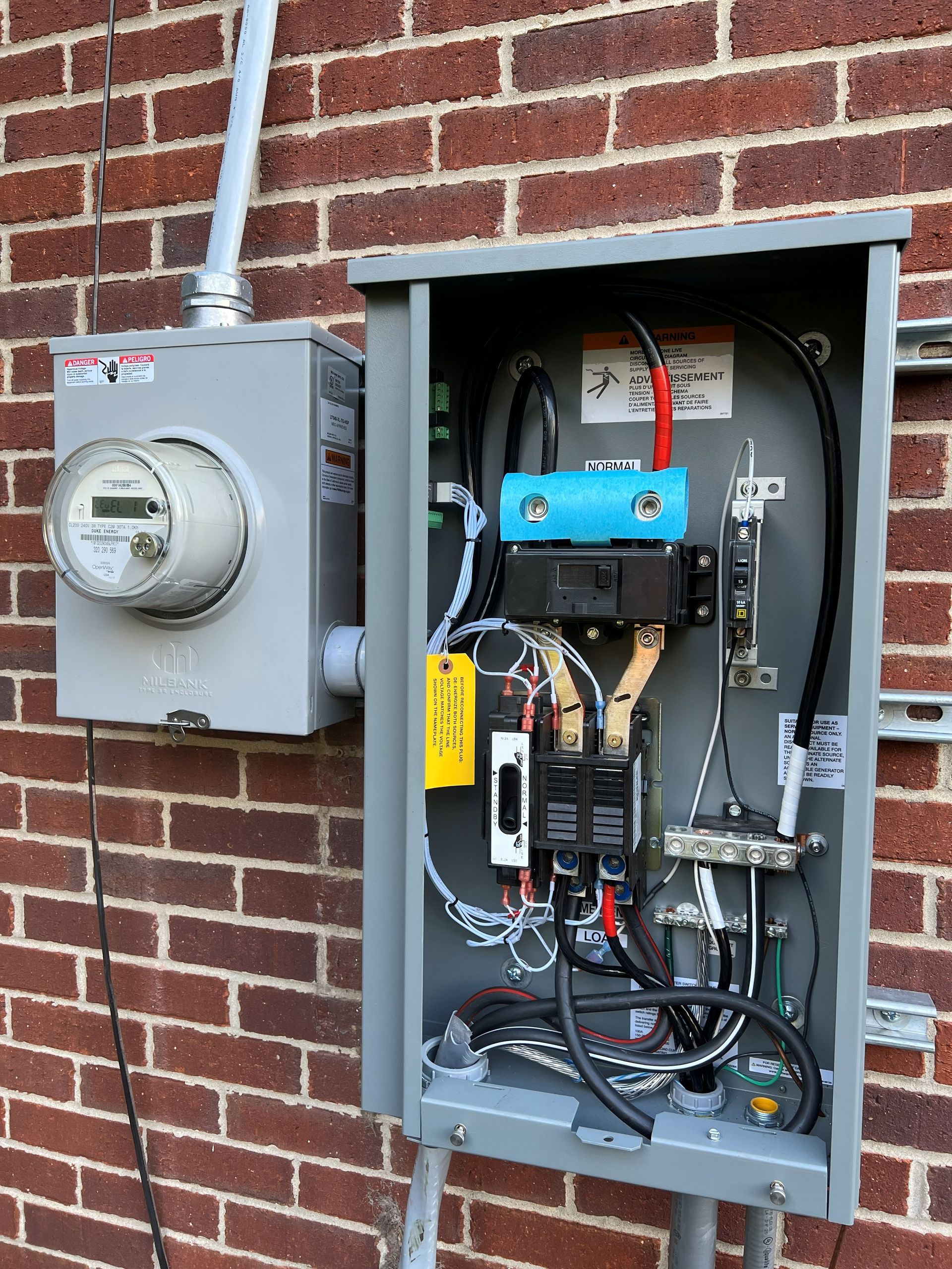 Electrical meter and breaker panel mounted on a brick wall. Gray metal components with wires.
