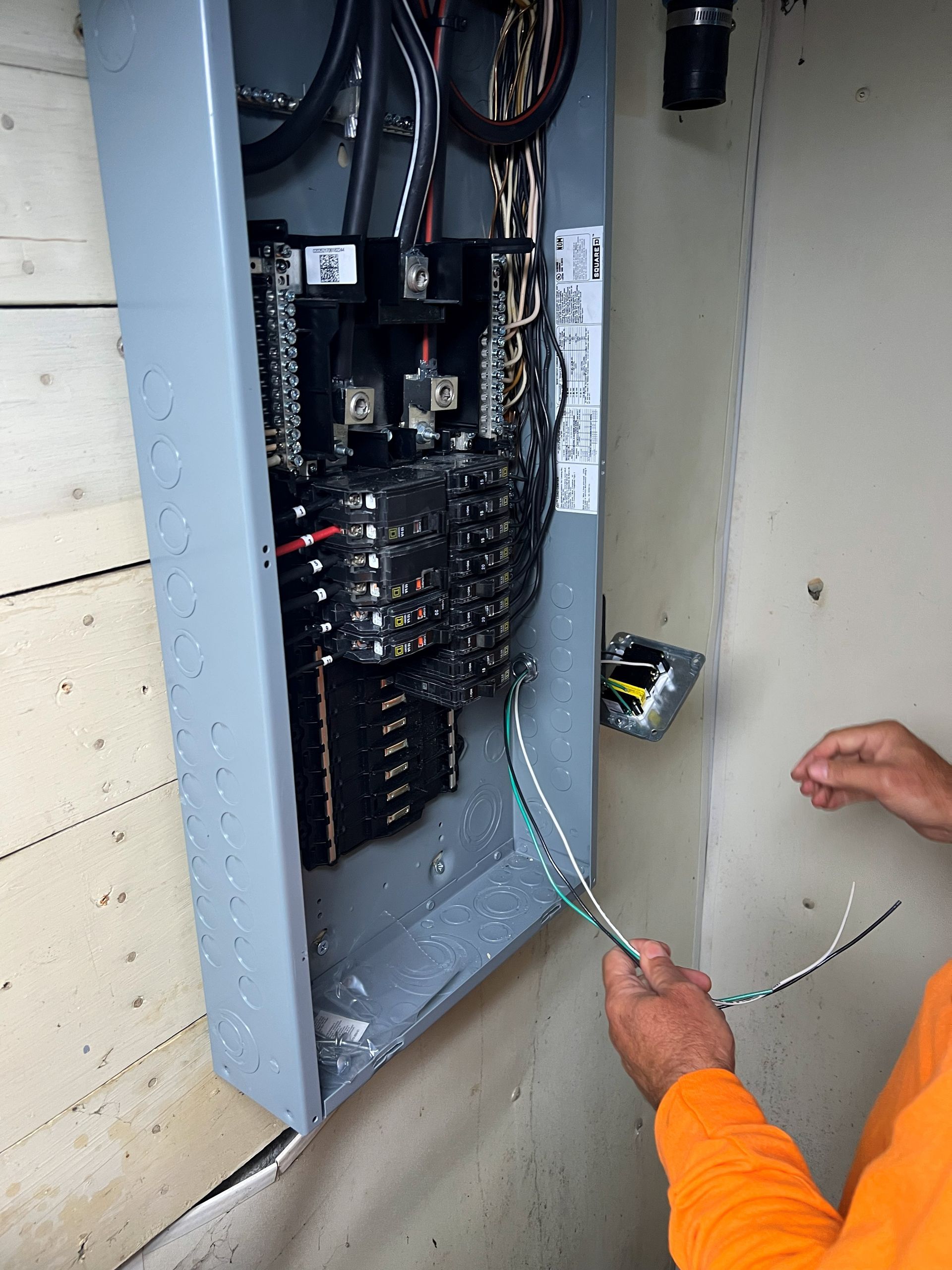 Electrician wiring an open electrical panel on a wall, holding wires. Gray panel, light wall.
