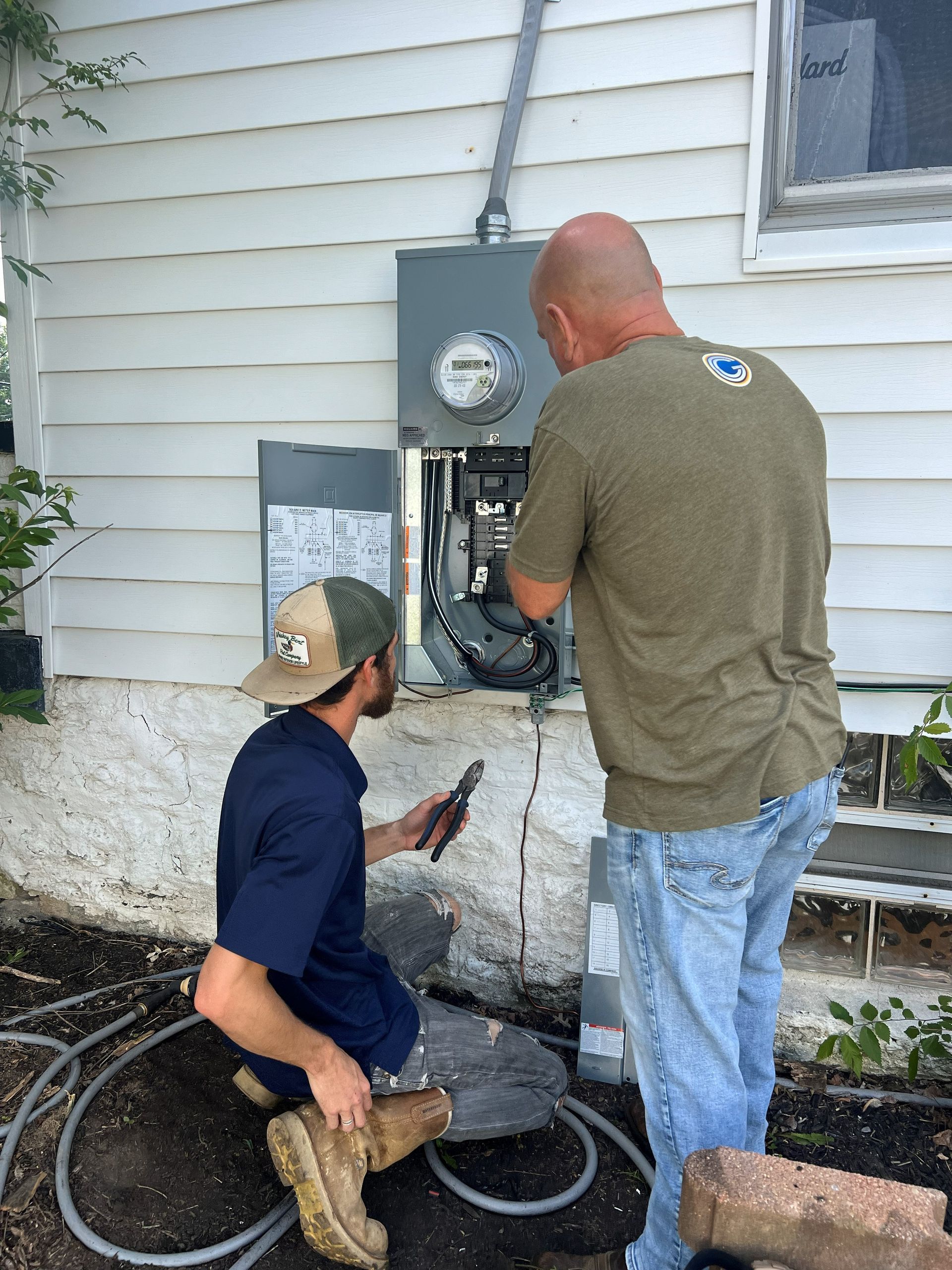 Two men working on an electrical panel outside a white house. One kneels with tools, the other stands, looking inside the panel.
