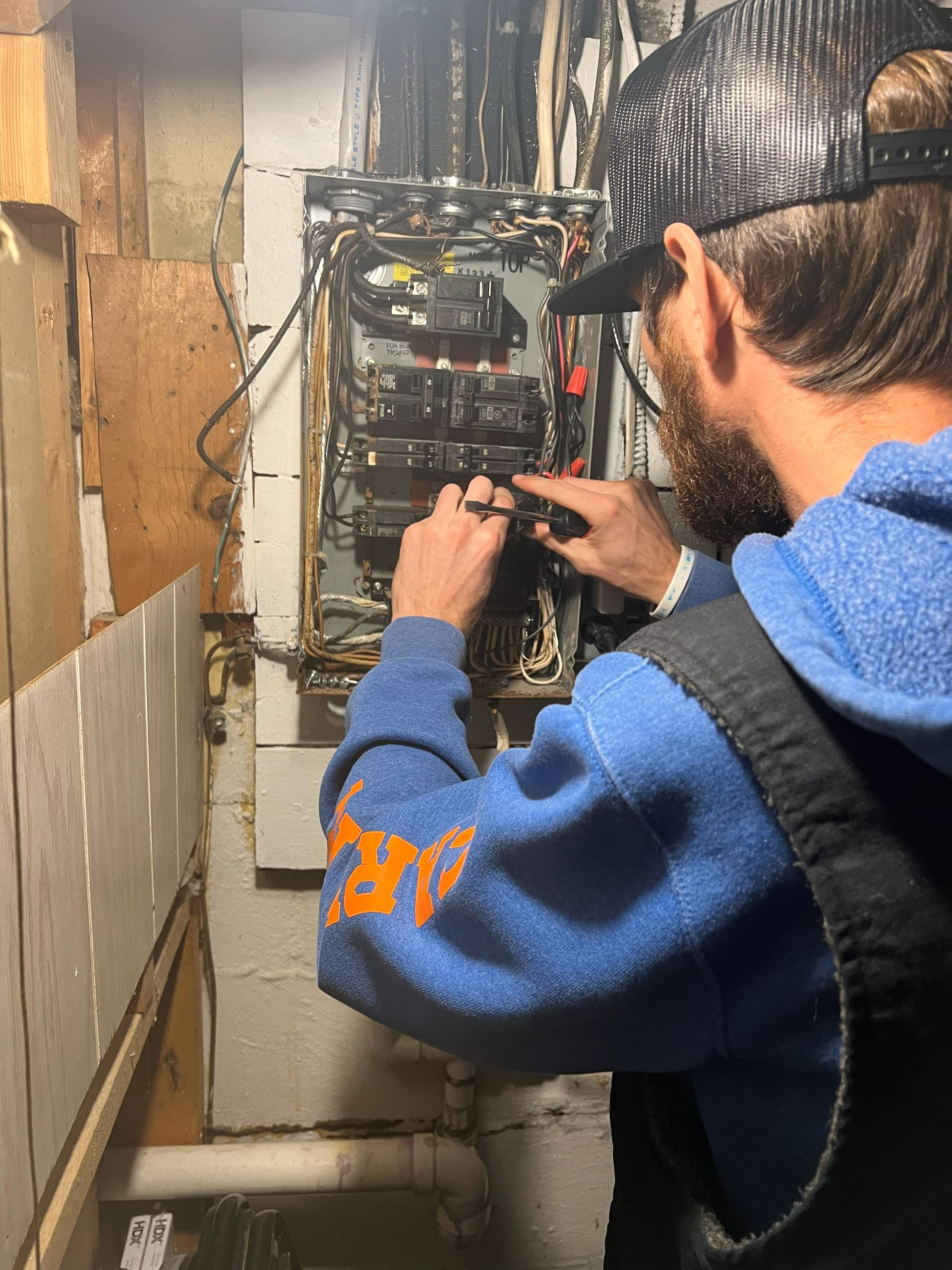 Person in blue hoodie working on an electrical panel in a room with exposed framing.