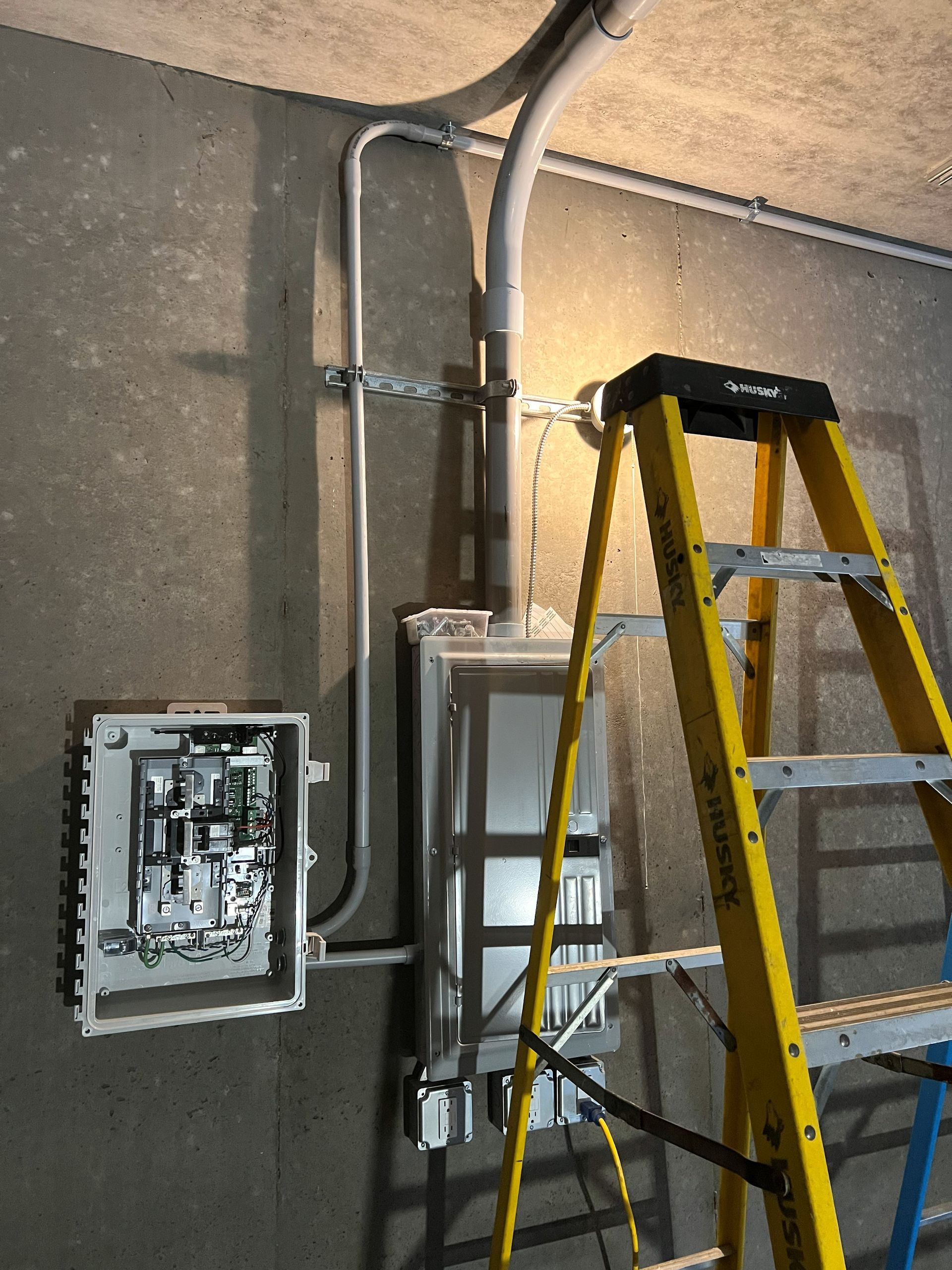 Electrical boxes on a concrete wall, with conduit and a yellow ladder nearby.