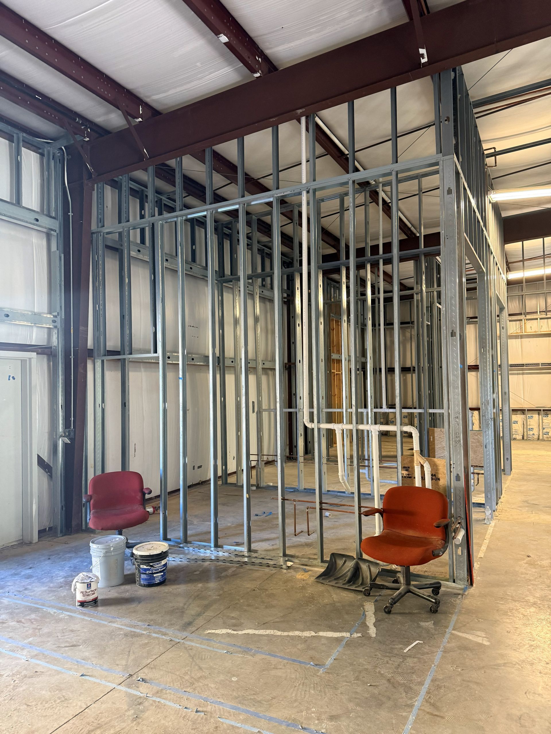 Metal framing of a room inside a large building. Two red office chairs and construction materials sit on the concrete floor.