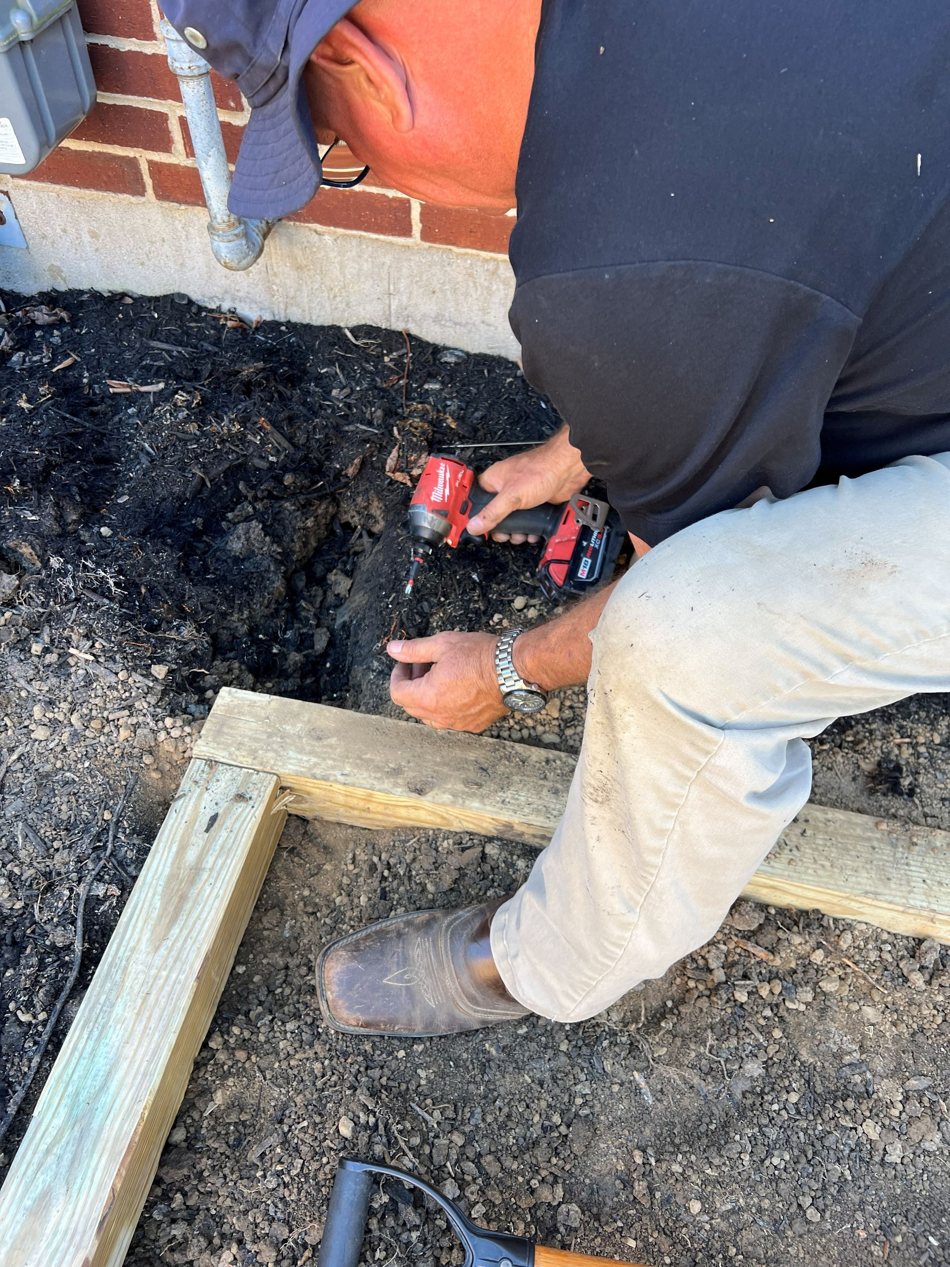 Person using a power drill to assemble wooden frame on a bed of soil next to a brick wall.