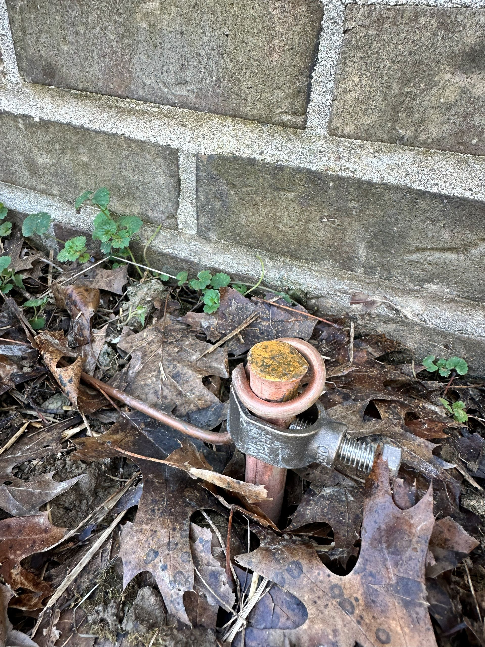 Copper grounding rod next to a brick wall, with a wire wrapped around its top. Surrounded by dirt and fallen leaves.