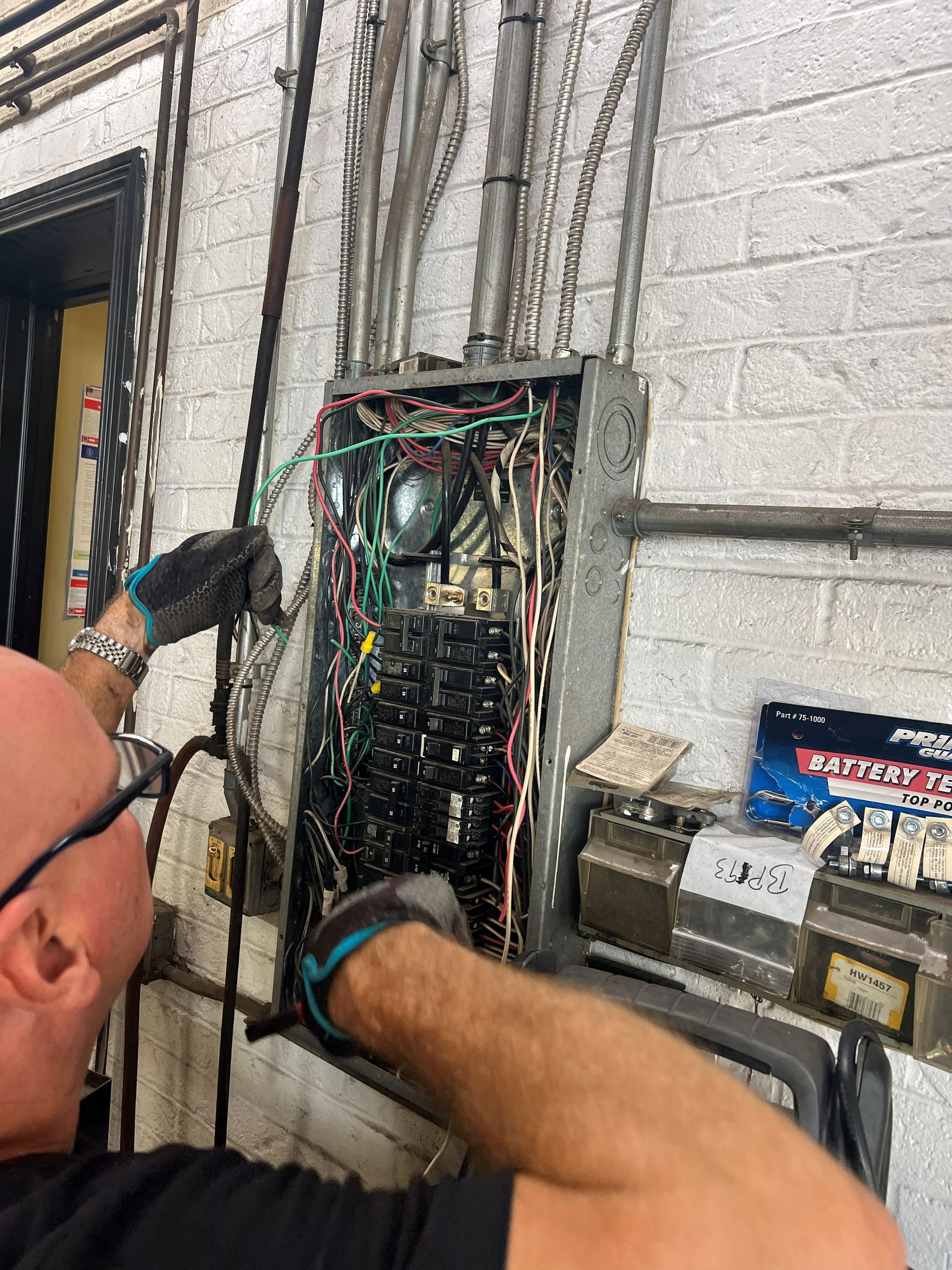 Electrician working on an open electrical panel, wires visible. Industrial setting, brick wall, wearing gloves.