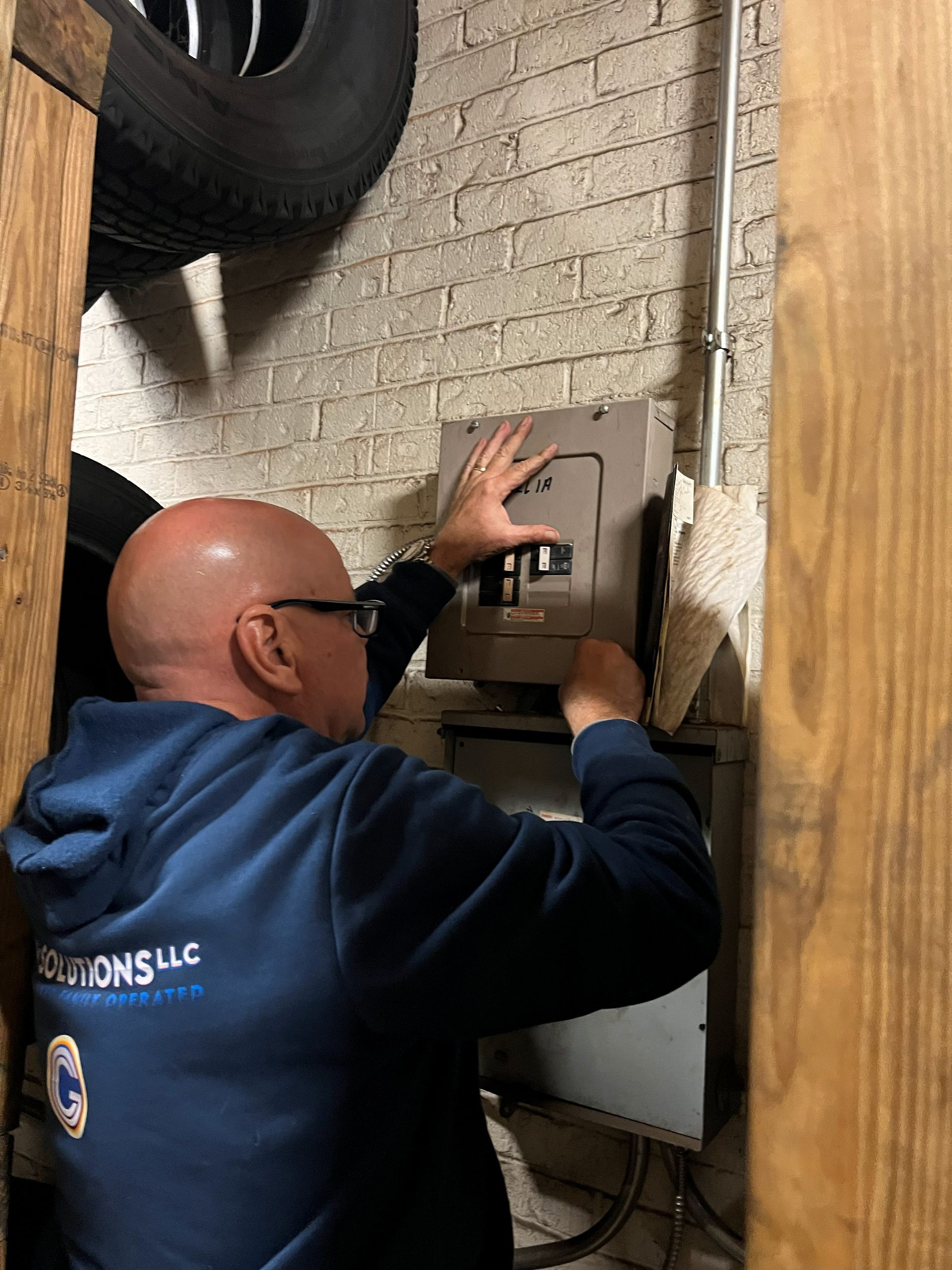 Man in blue hoodie working on electrical panel mounted on brick wall.