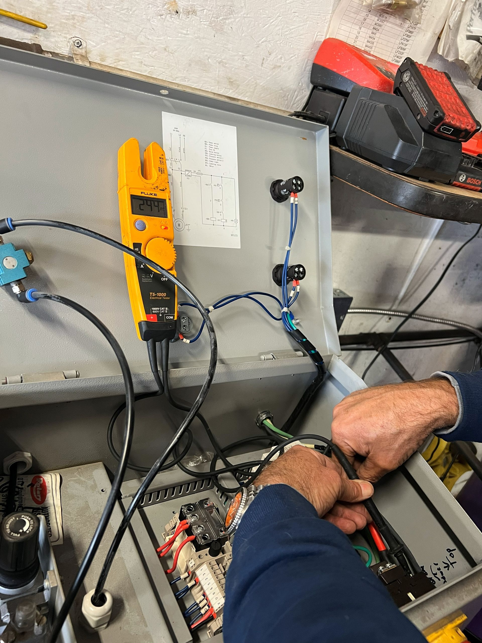 Person using a multimeter to test electrical components inside a gray control panel.