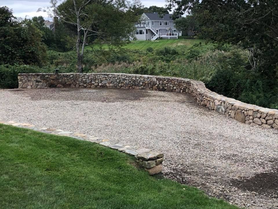 A stone wall surrounds a gravel driveway with a house in the background.