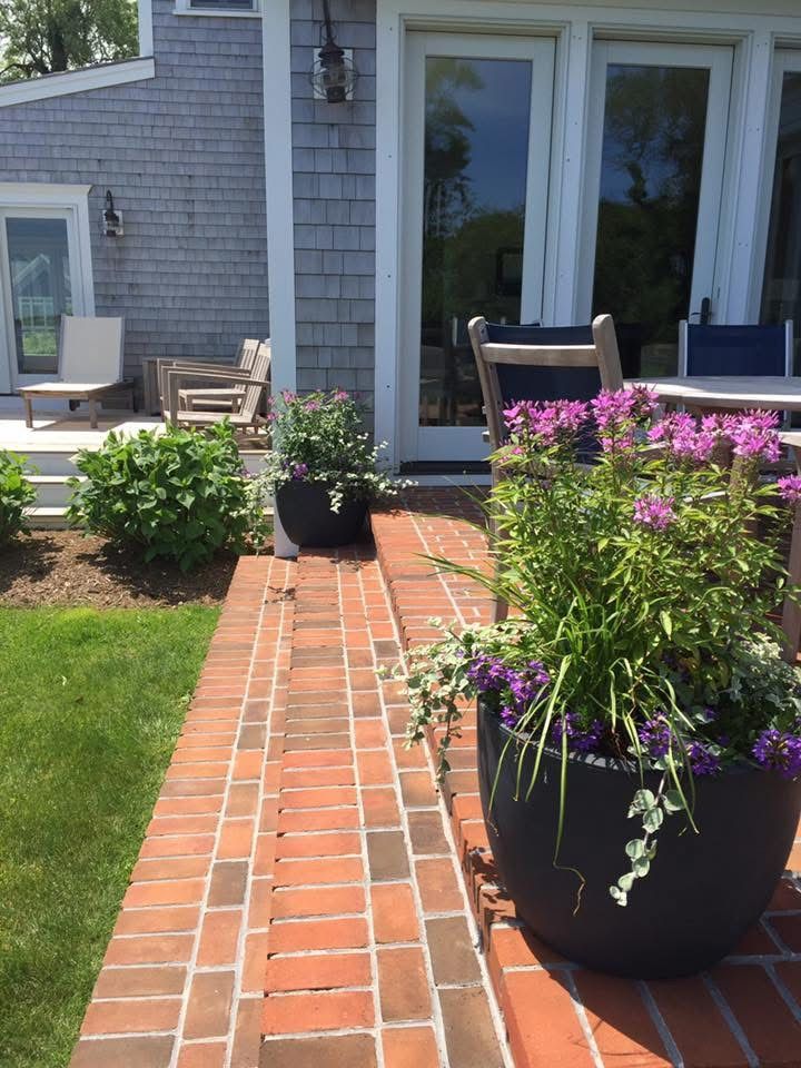 A brick walkway leading to a patio with purple flowers in pots