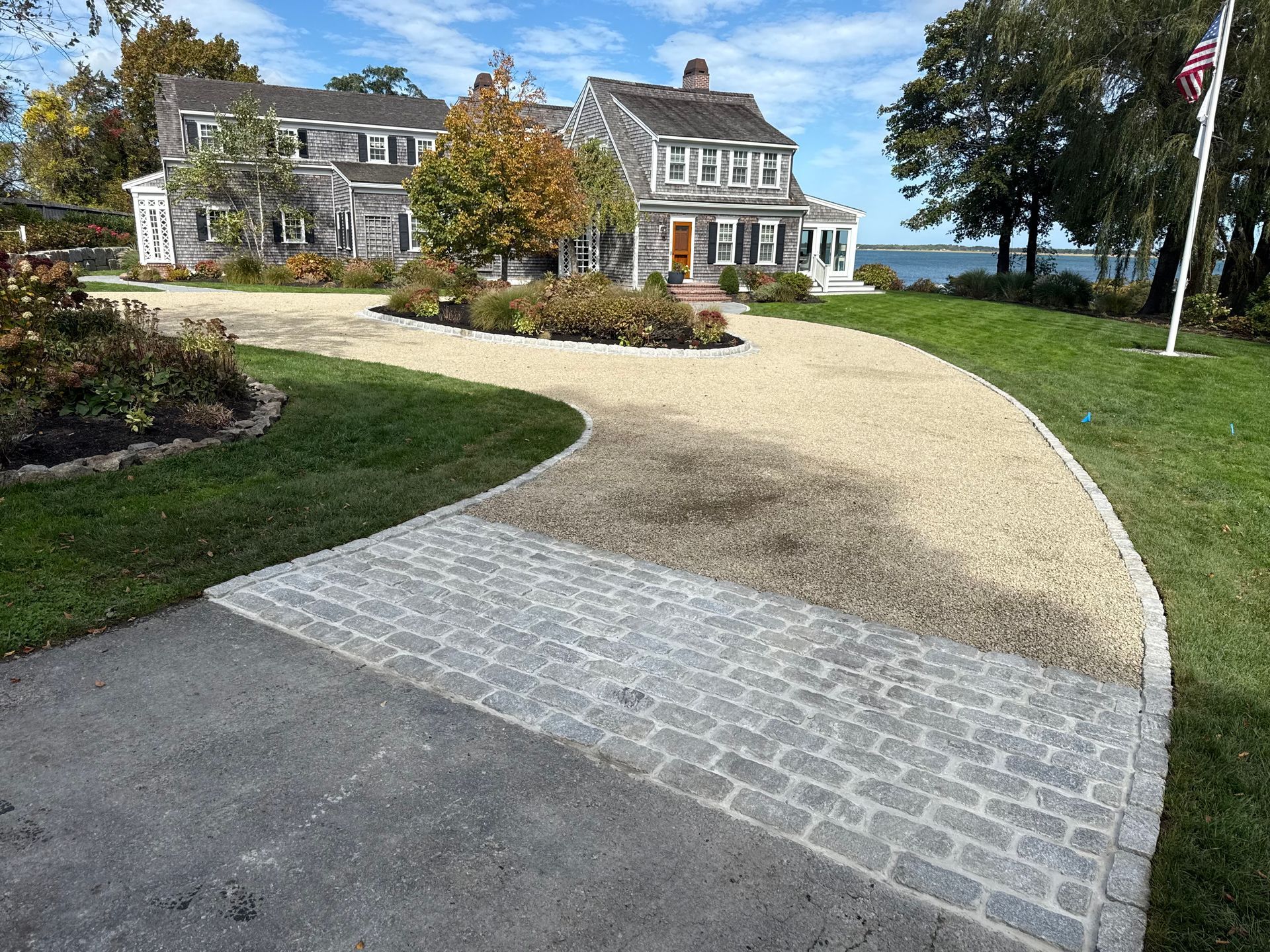 Driveway leads to a two-story gray house, with cobblestone and gravel path, and green lawn.