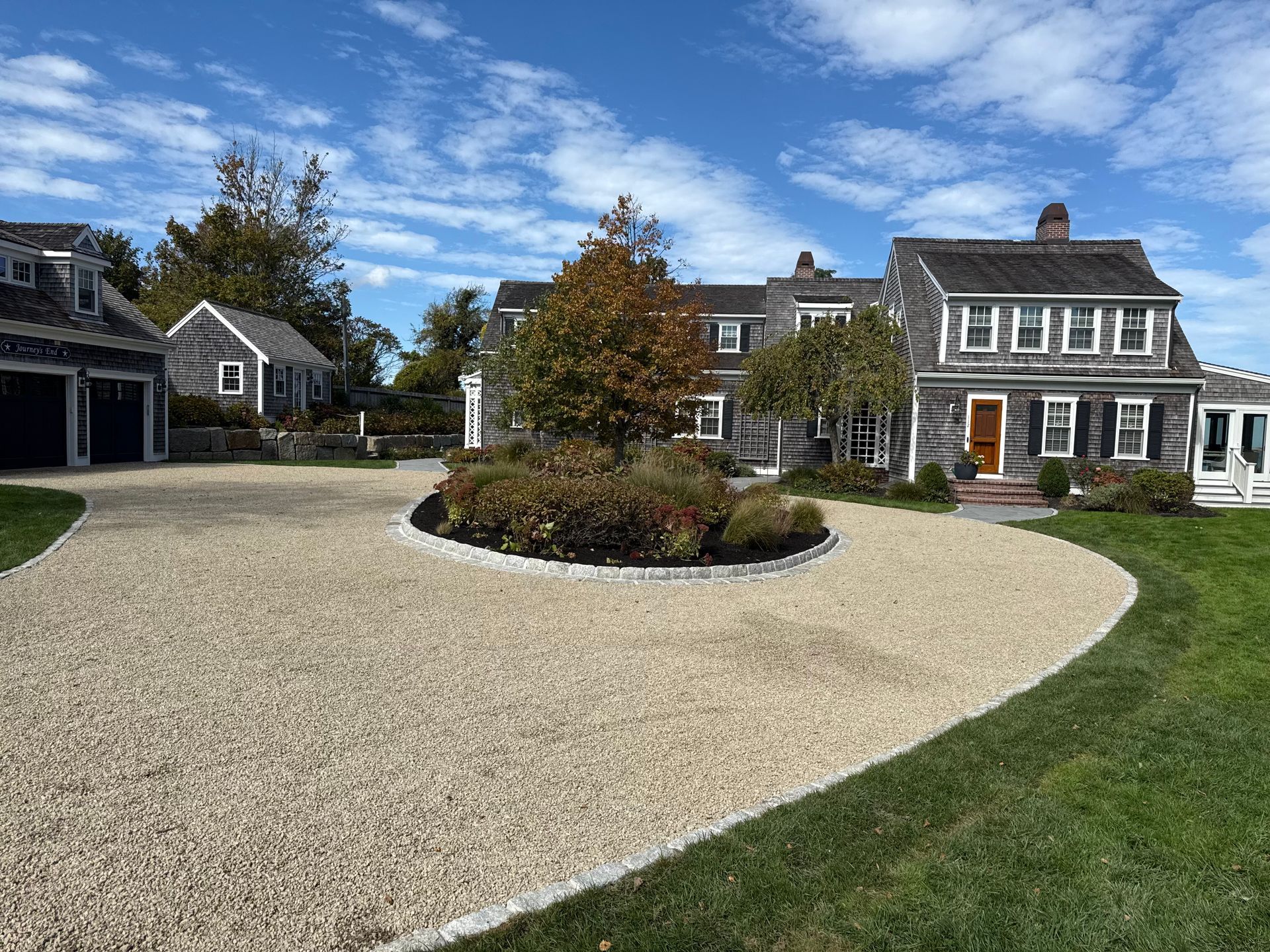 Gravel driveway leads to a weathered gray house with landscaping, blue sky.