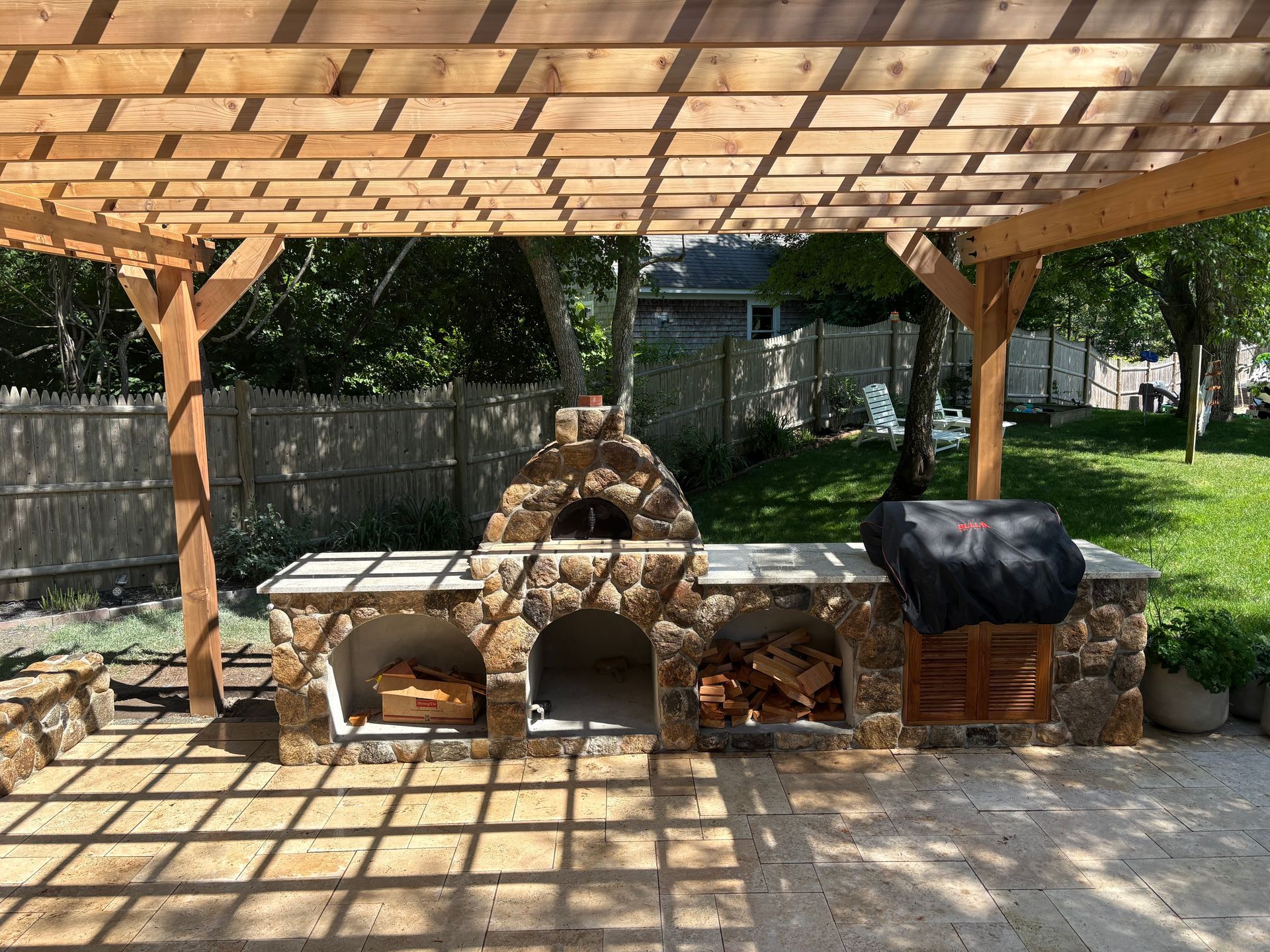 Outdoor stone pizza oven under a wooden pergola with a grill and firewood in a backyard patio
