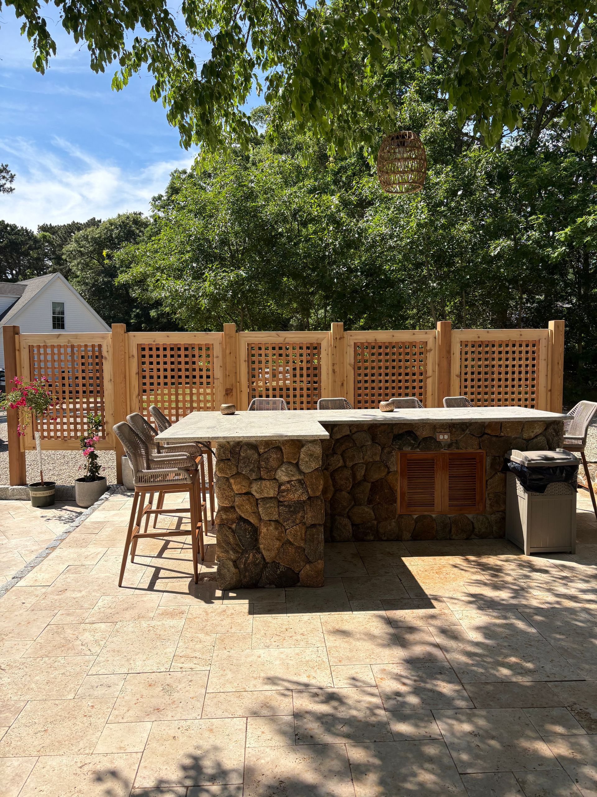 Outdoor stone patio with built-in grill and sink, surrounded by wooden lattice fencing under trees.
