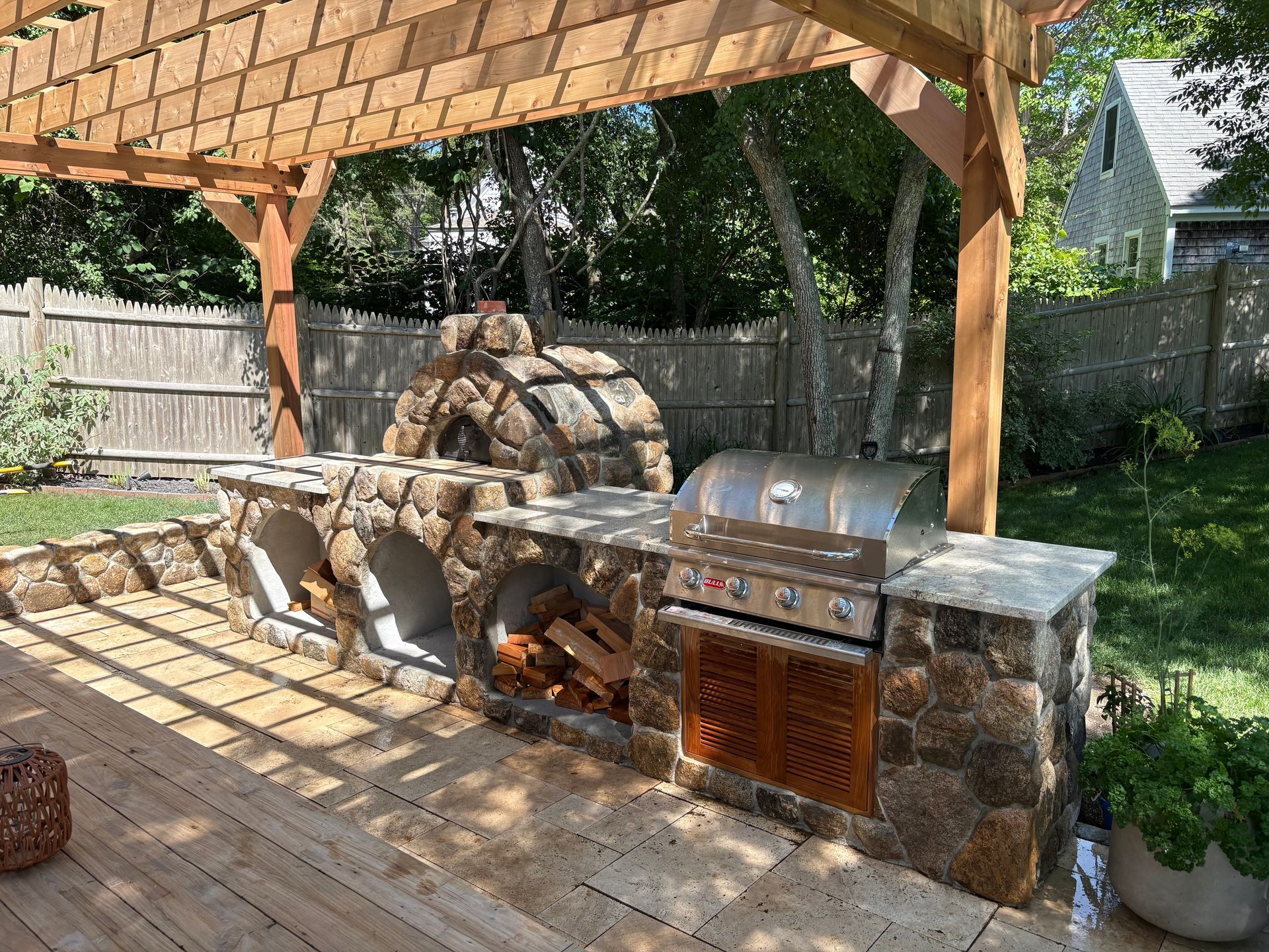 Backyard outdoor kitchen under a pergola with stone grill, sink, and storage cabinets