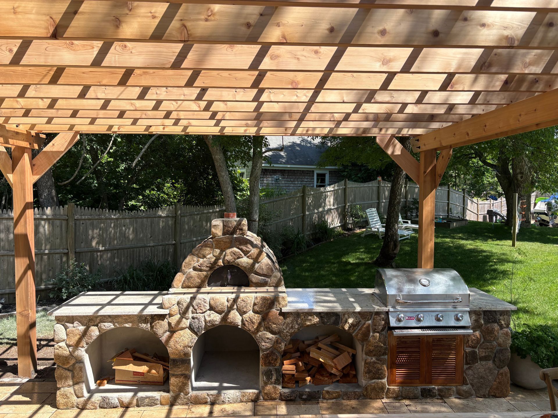 Outdoor stone barbecue and pizza oven under a wooden gazebo in a backyard