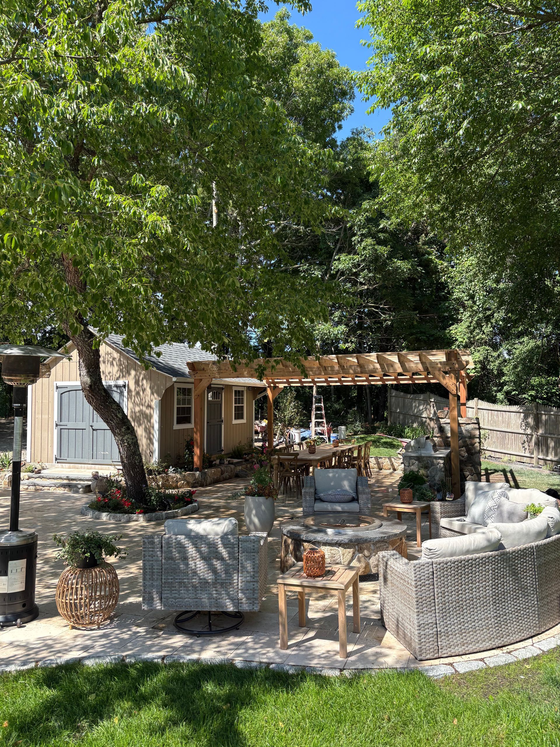 Shaded outdoor patio with wicker seating, tables, and a wooden pergola under leafy trees