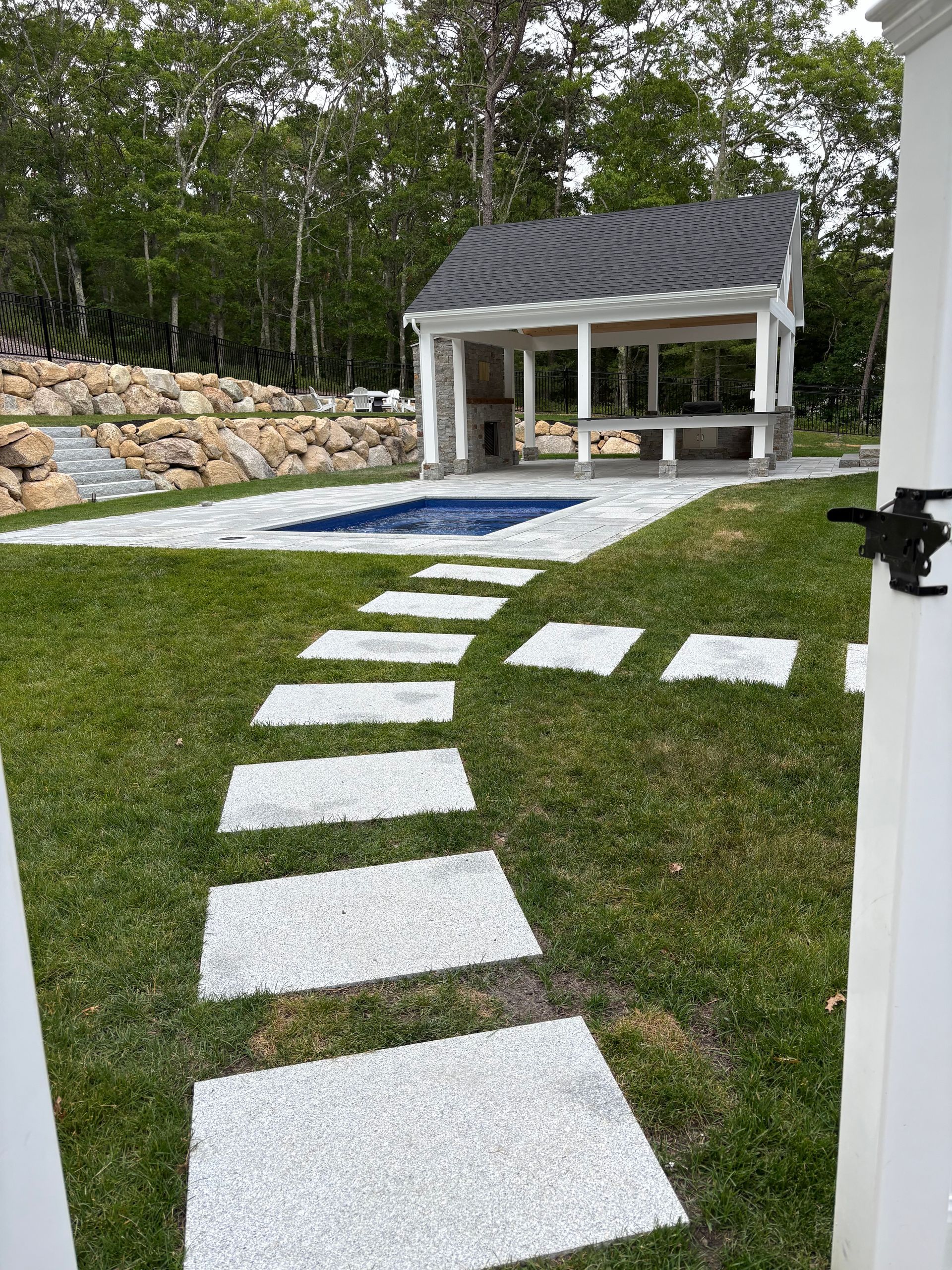 Curved stepping stones through a green lawn toward a small white gazebo beside a pool and stone wall