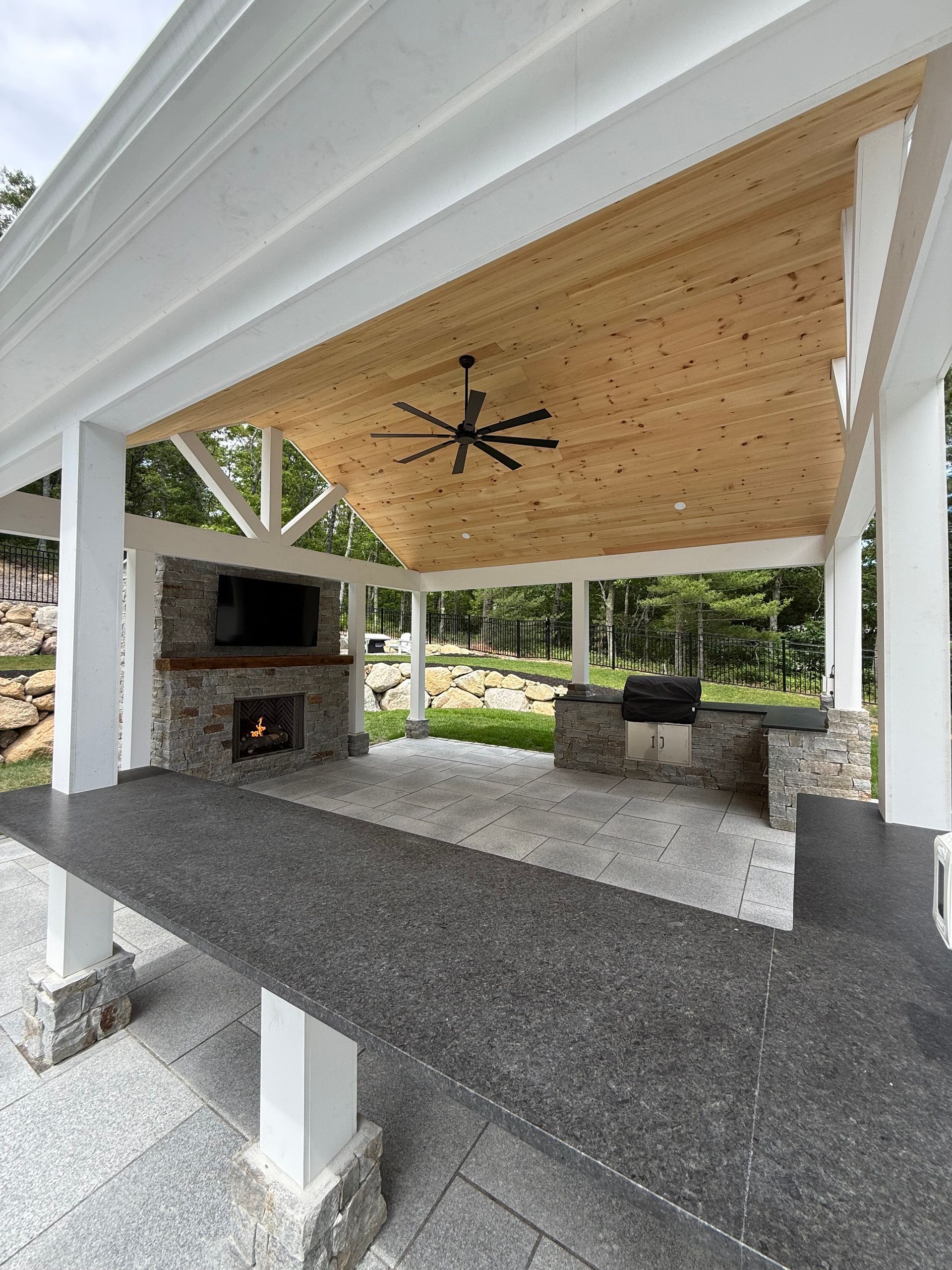 Covered outdoor patio with stone fireplace, black ceiling fan, and white support columns under a wood roof