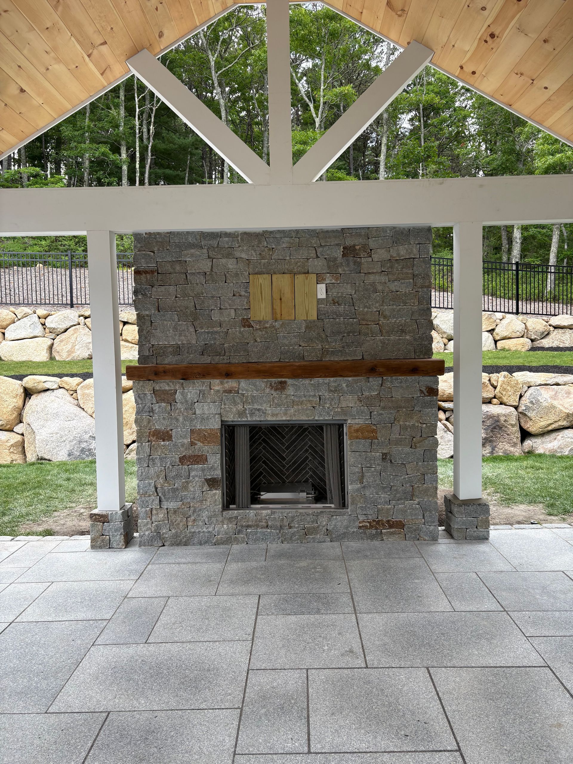 Stone outdoor fireplace under a wooden pavilion, with gray stone floor and trees in the background